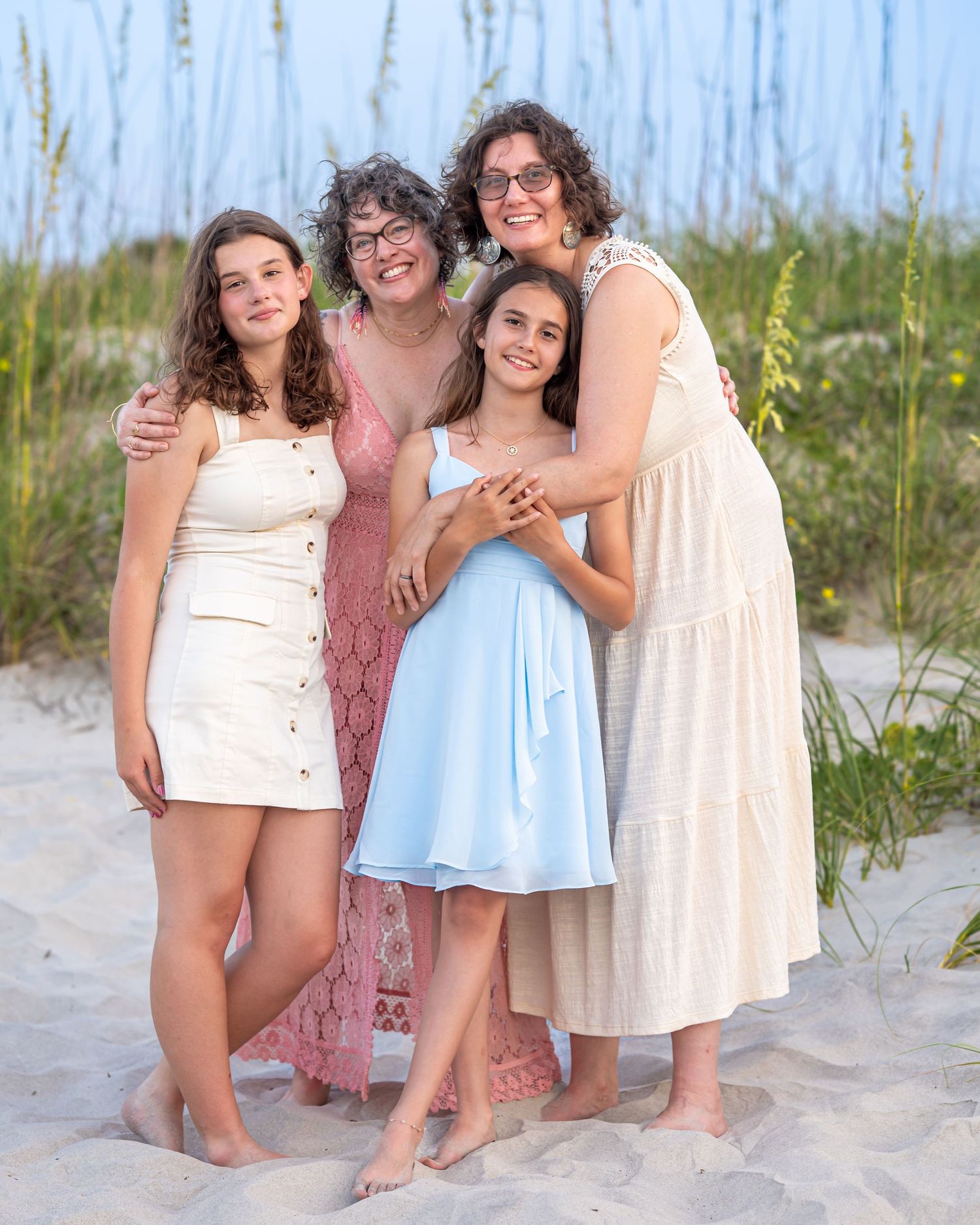 Two moms and their daughters posing together on a sandy beach, wearing summer dresses, smiling warmly near sea oats.