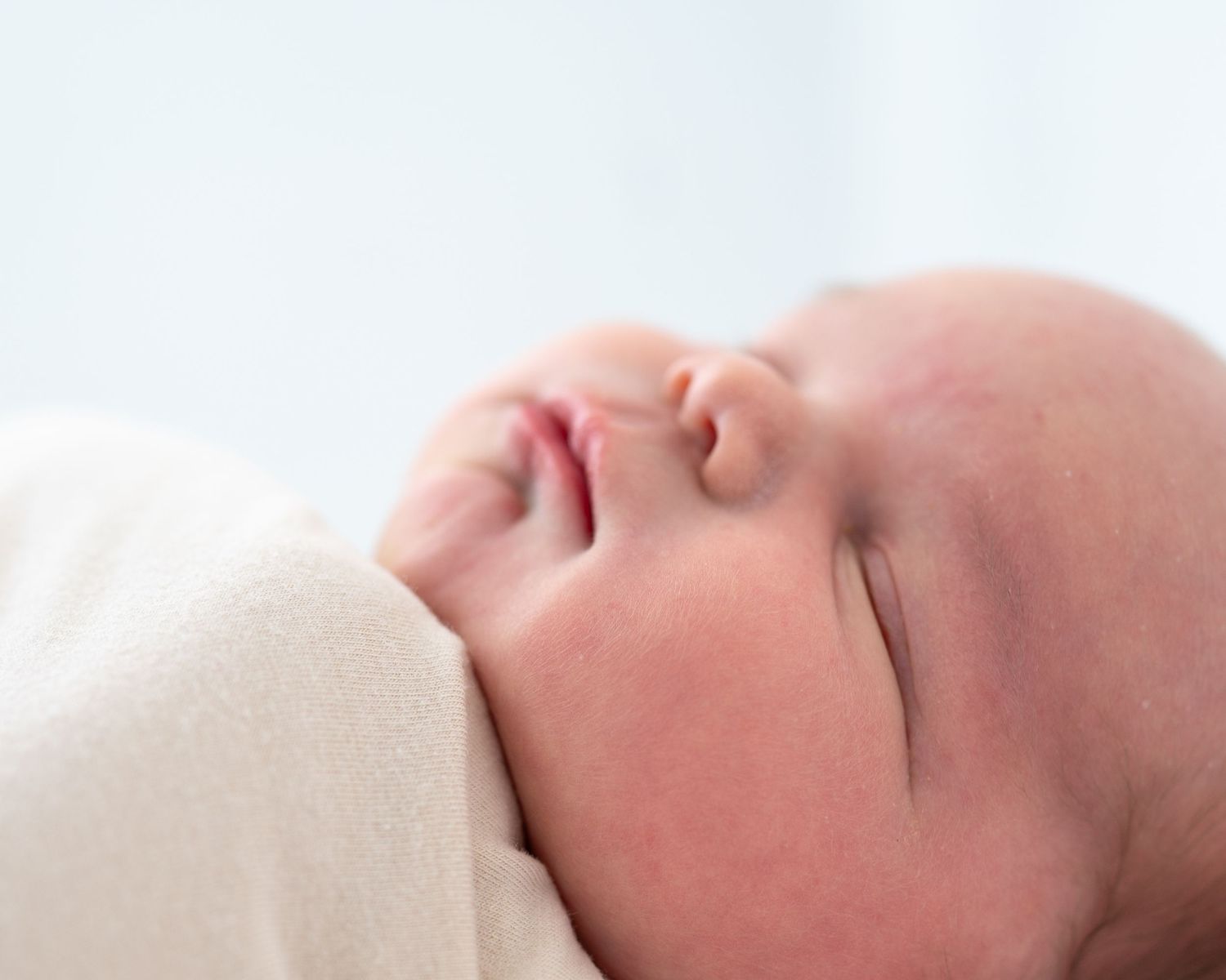 Peaceful newborn baby sleeping soundly, swaddled in a soft cream blanket with delicate features in a serene close-up.