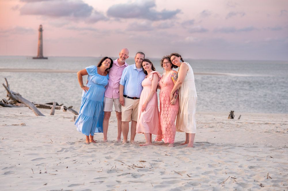 Family of six poses on sandy beach at sunset near a lighthouse, wearing pastel outfits in pink and blue tones.