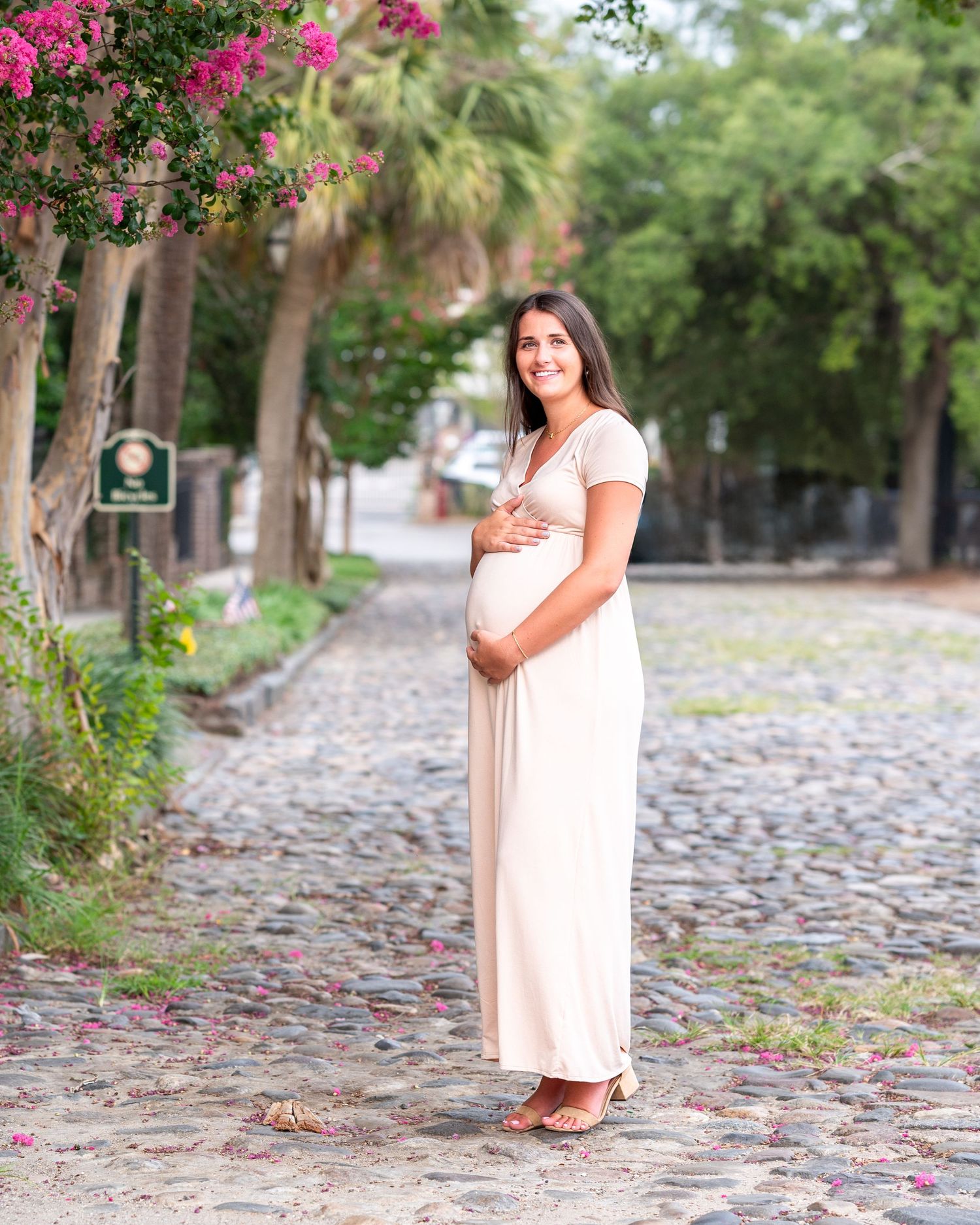 Pregnant woman in white dress holds her bump while standing on cobblestone street