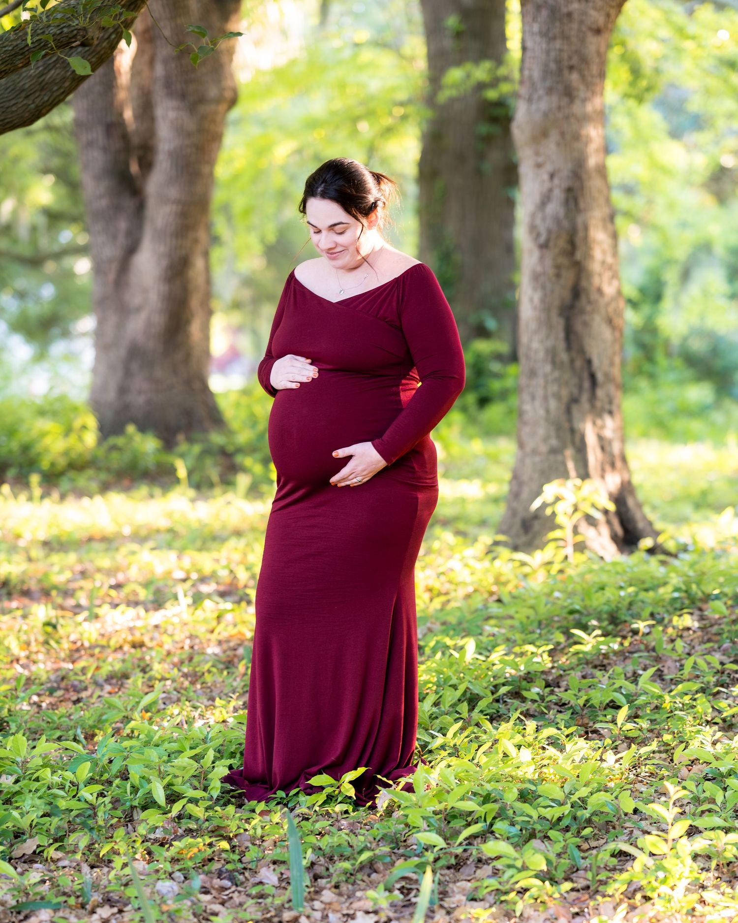 Lucy stands in a burgundy dress in a field with trees, she looks down at and embraces her pregnant belly.