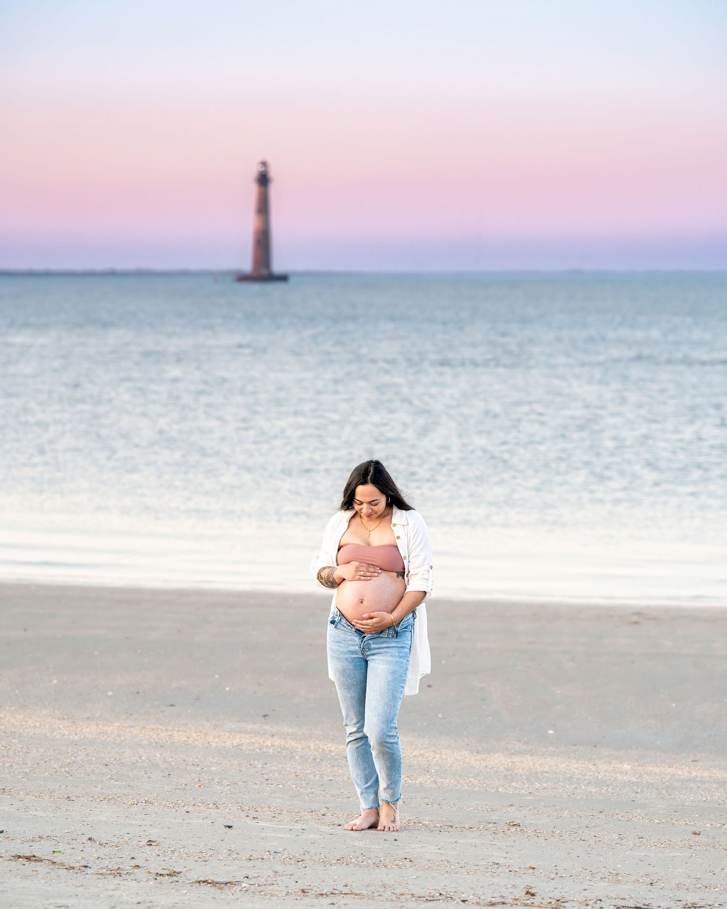 Person walking on a sandy beach at sunset with Morris Island Lighthouse visible in the background off the South Carolina coast.