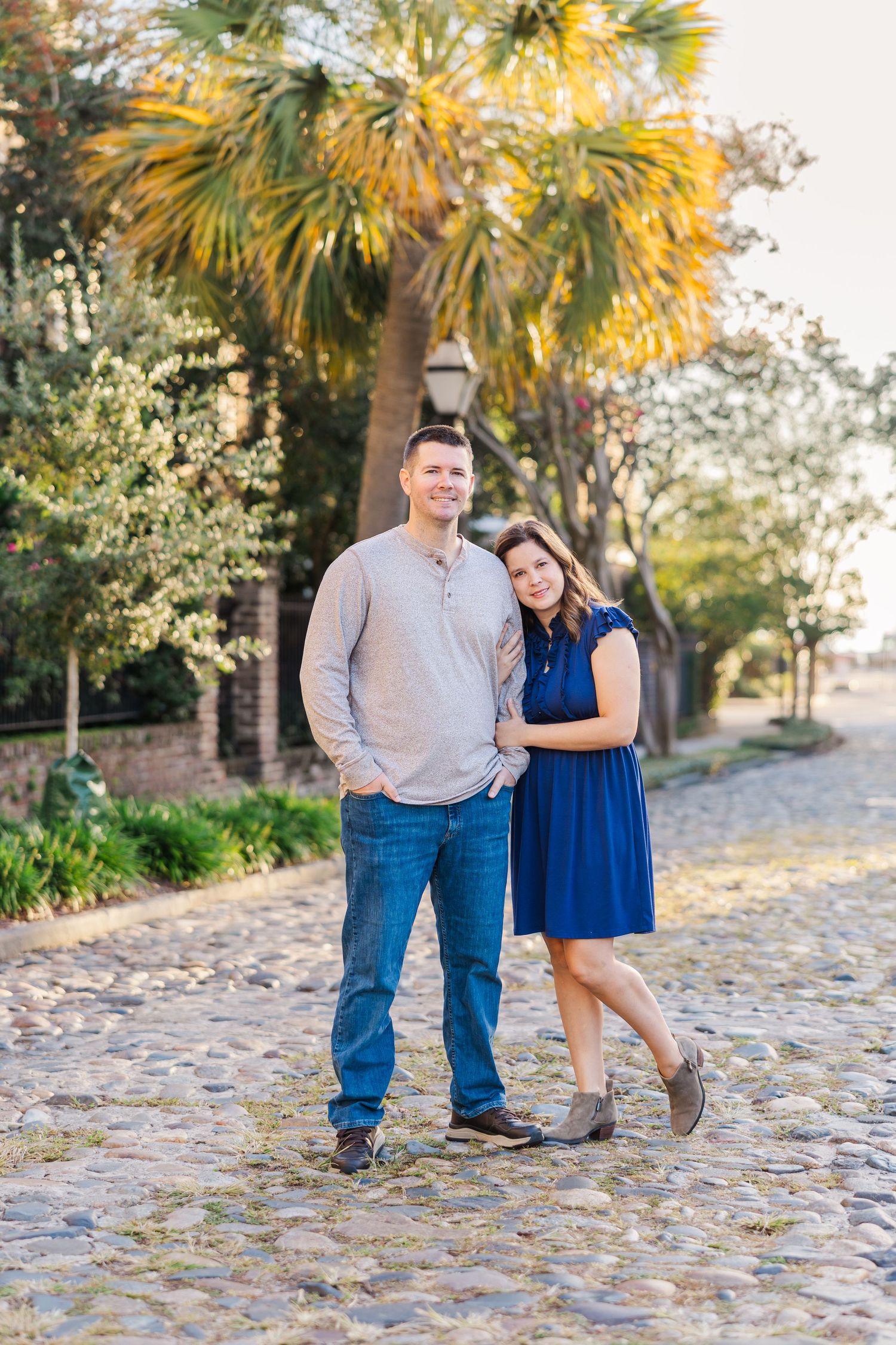 A couple stands together on a cobblestone path under palm trees in Charleston's historic district on a sunny afternoon.