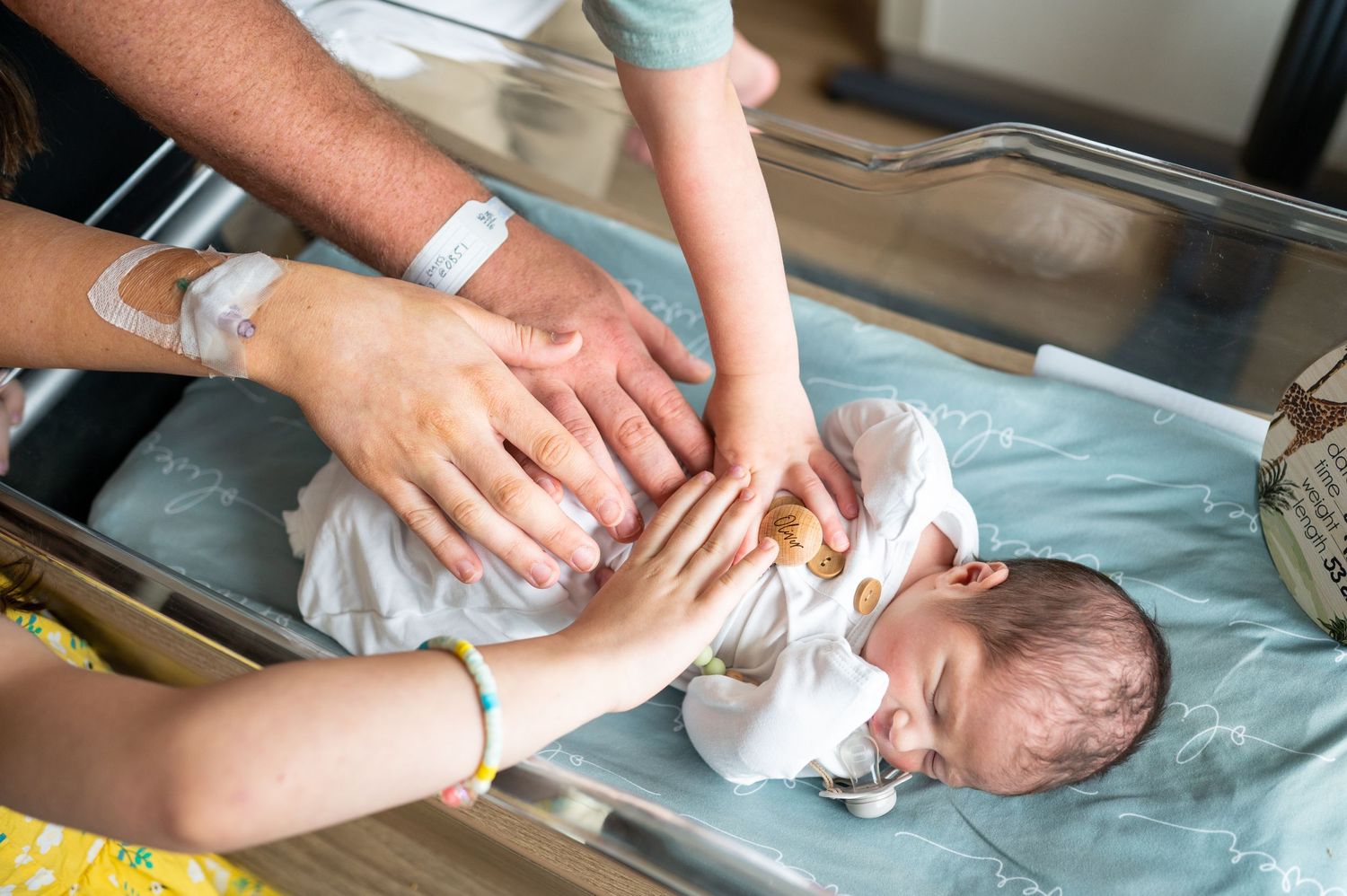 A newborn baby boy lays in his hospital bassinet with his mom's, dad's, sister's and brother's hands on his belly.