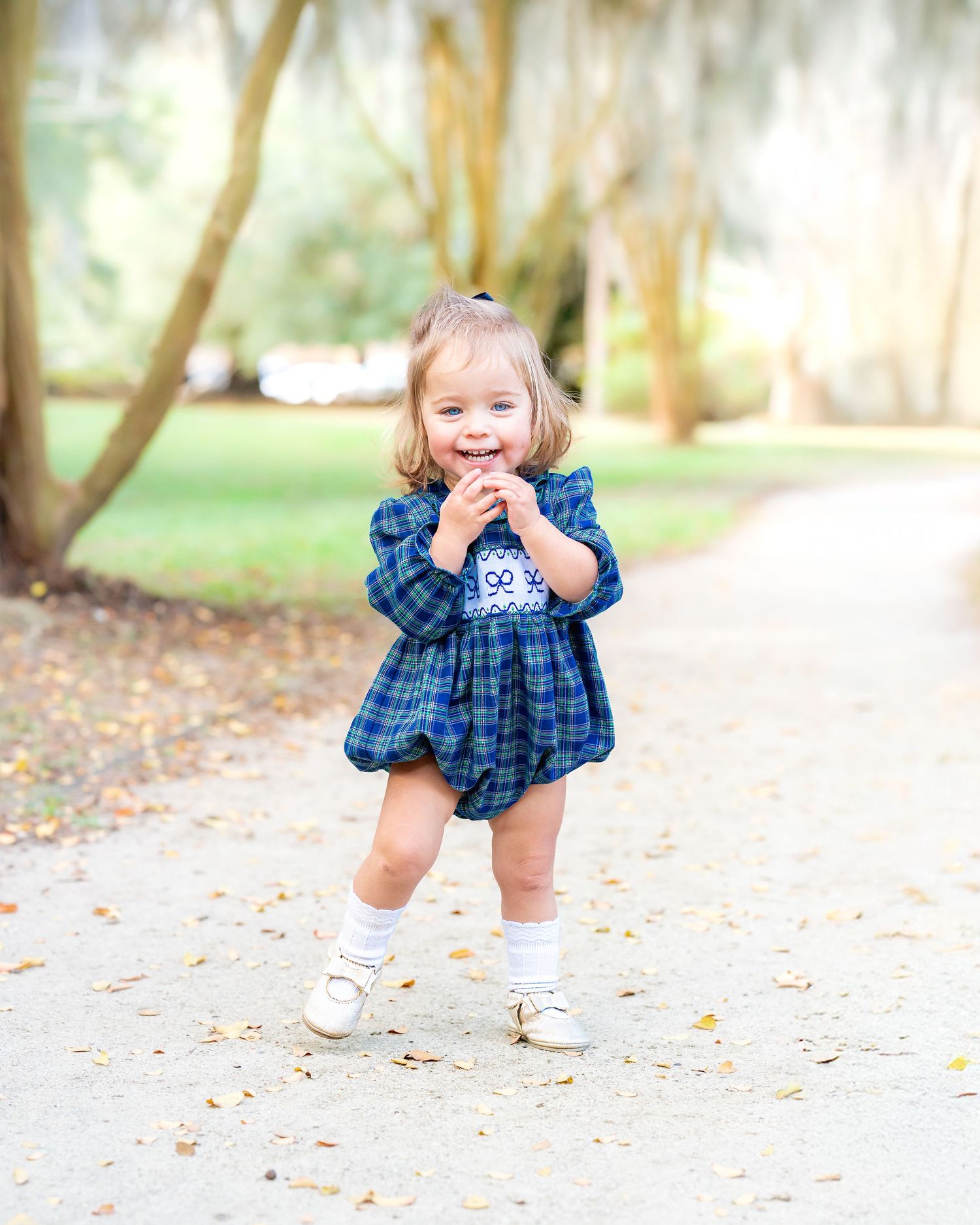 Young child in blue plaid dress playing and laughing outdoors on a path with fallen leaves.