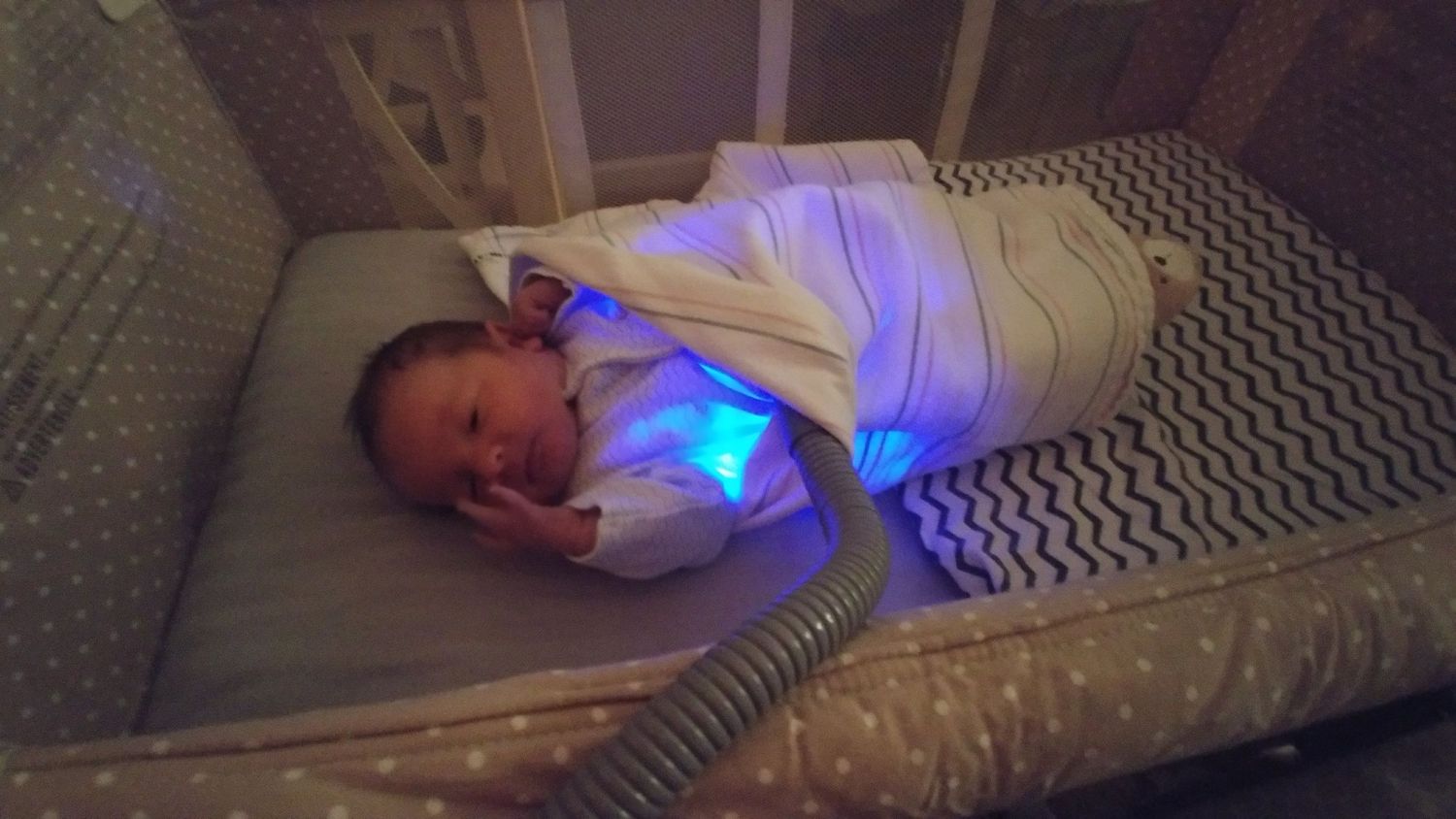 A sleeping infant in a crib illuminated by a bill lights rests on a chevron patterned mattress.