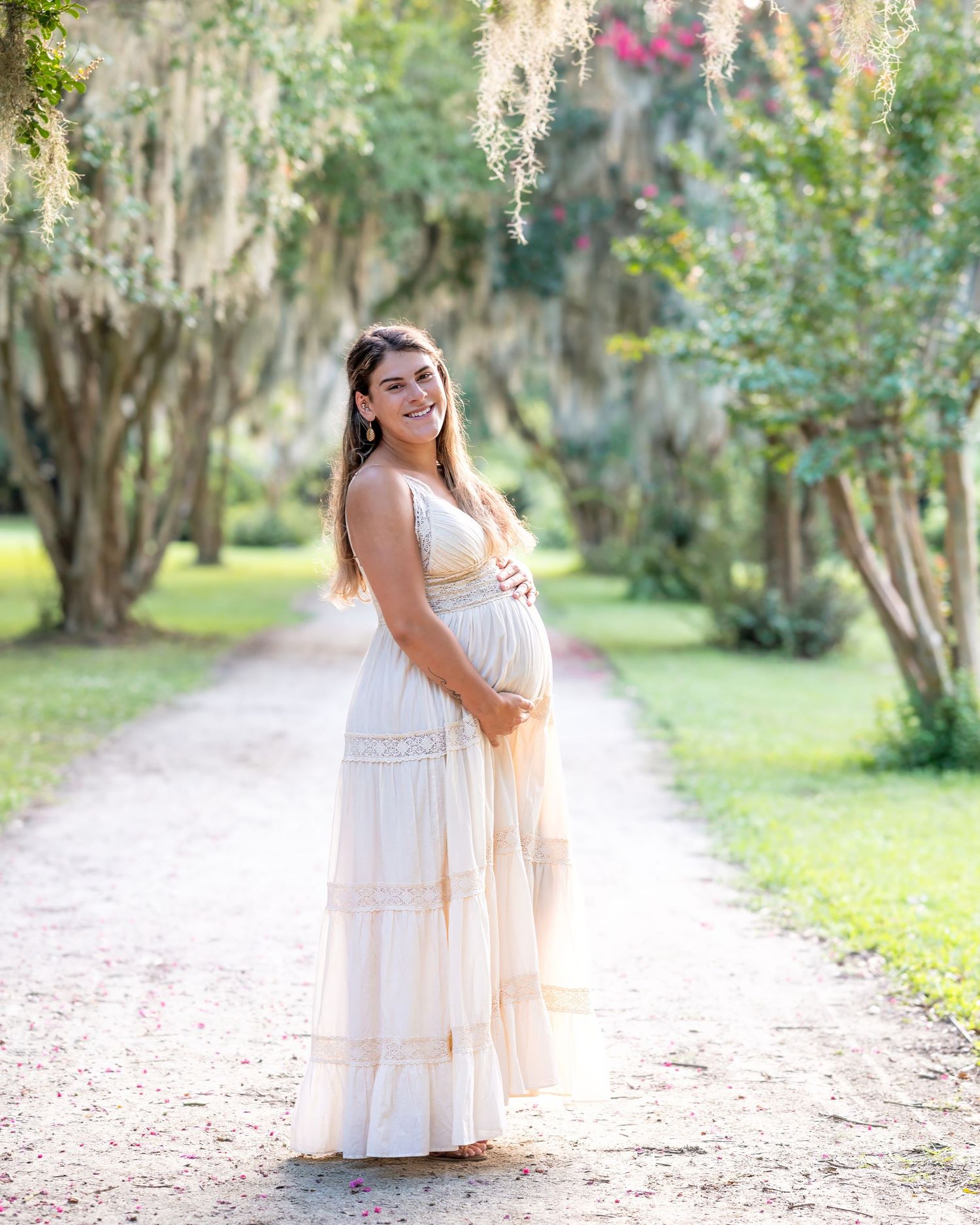 Lola stands laughing and holding her pregnant stomach on a path lined with trees draped in Spanish moss.