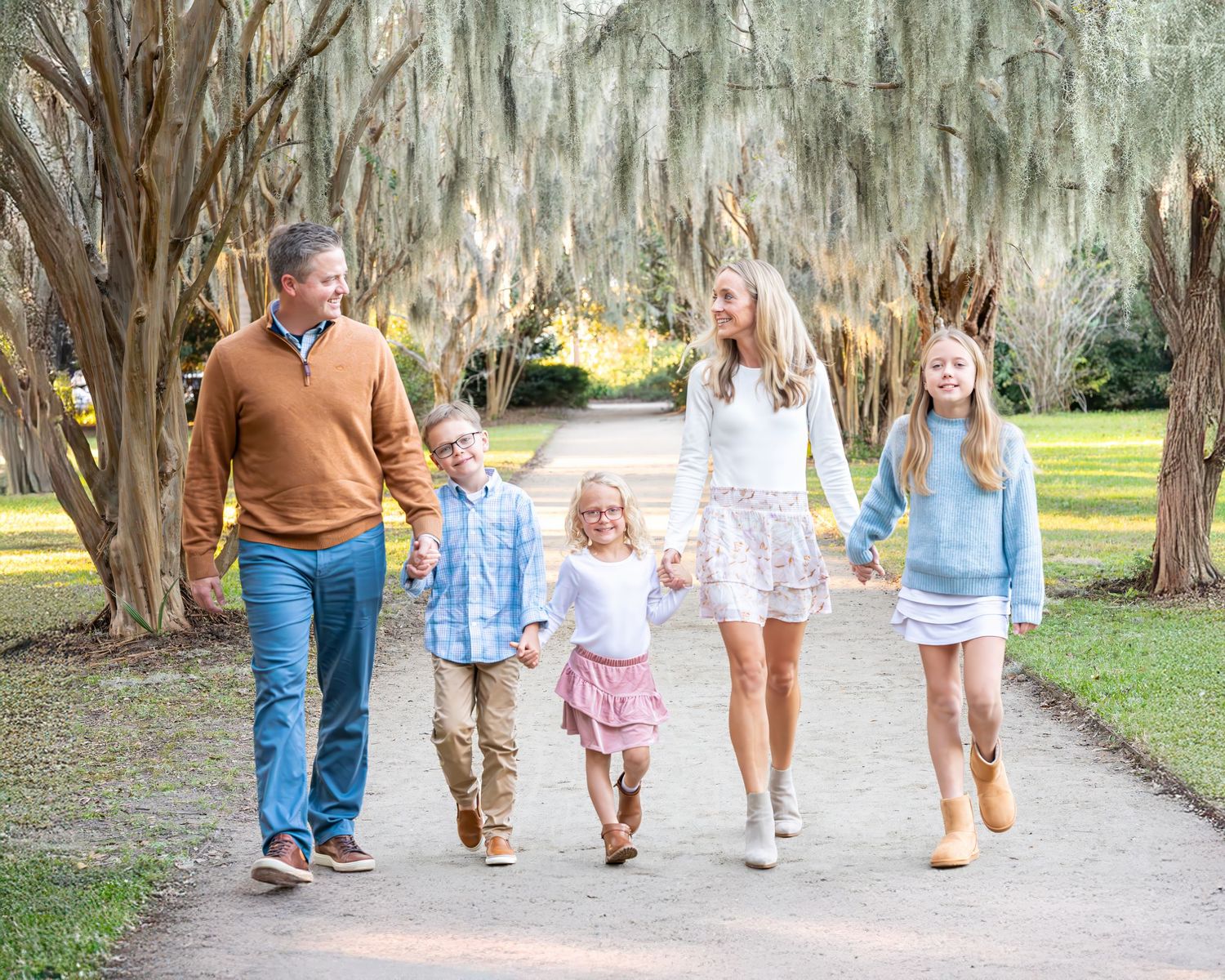A happy family walks together on a path under moss-draped trees in a scenic Southern garden setting.