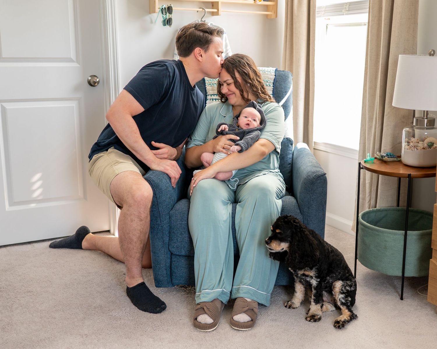 New parents share a tender kiss over their newborn baby in a nursery chair, with their black and white dog nearby.
