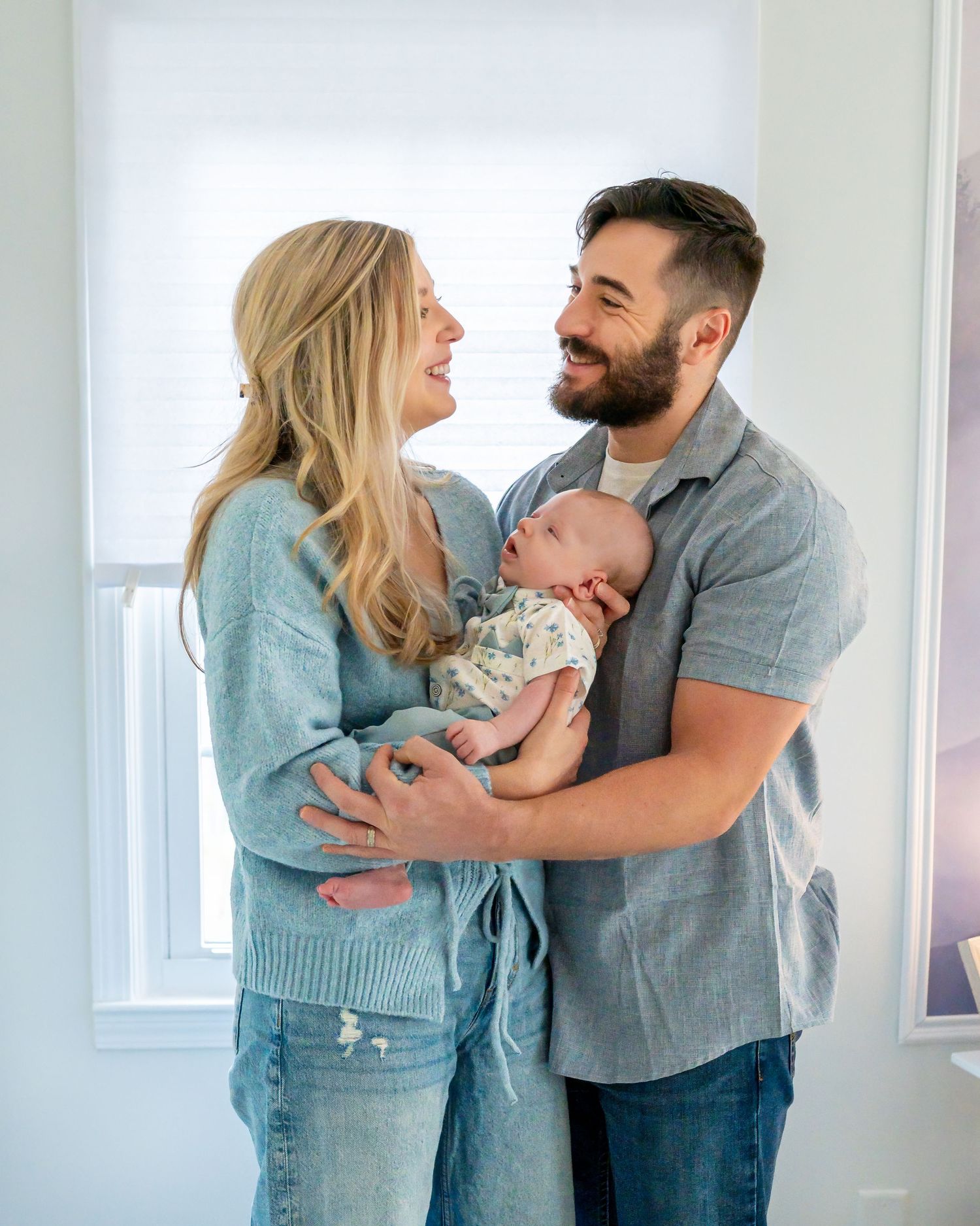 Happy couple in casual attire sharing a tender moment while holding their baby in a bright room by a window.