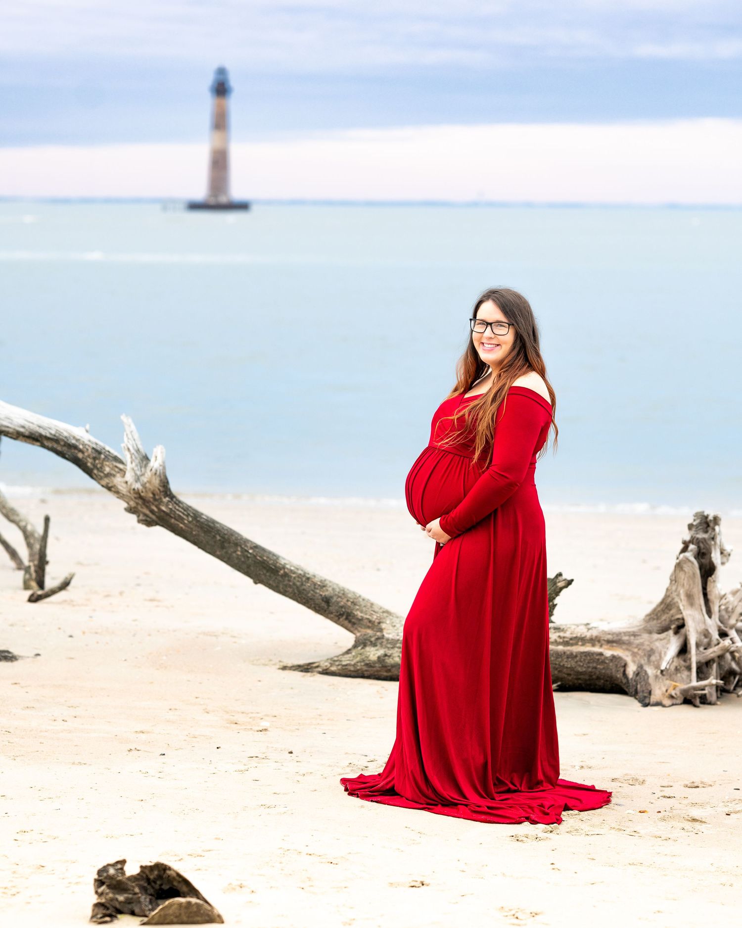 Woman in flowing red gown stands on beach with lighthouse and driftwood in background.