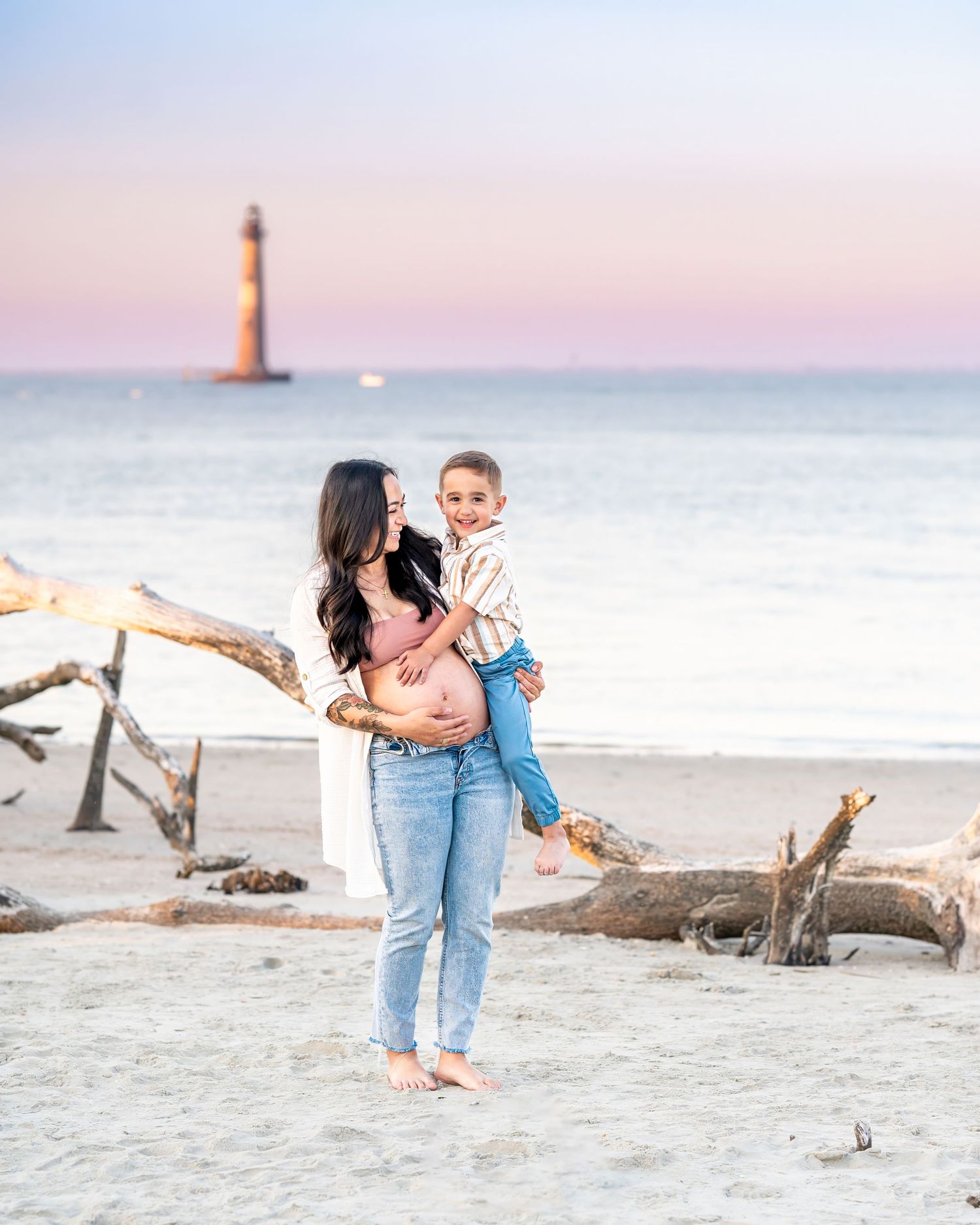 A sunset beach scene with a lighthouse in the background and driftwood on the sand.