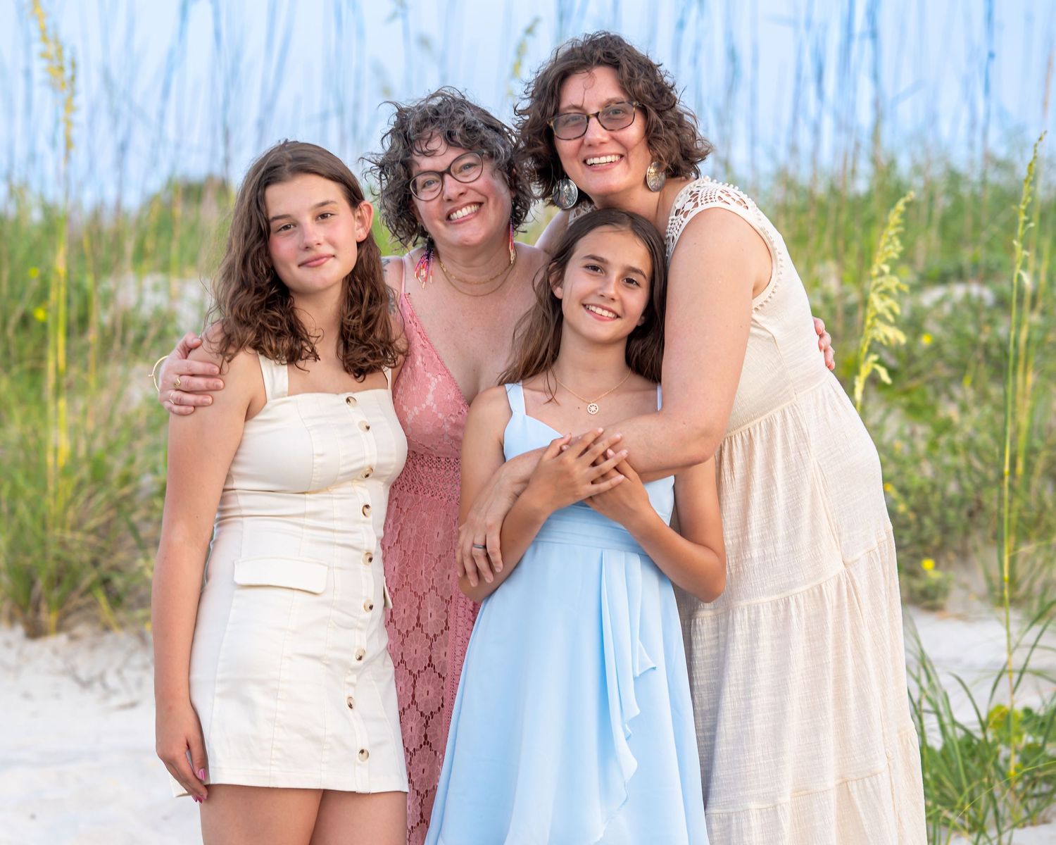Four women smiling together on a sandy beach with sea oats, wearing summer dresses in white, blue, and pink.