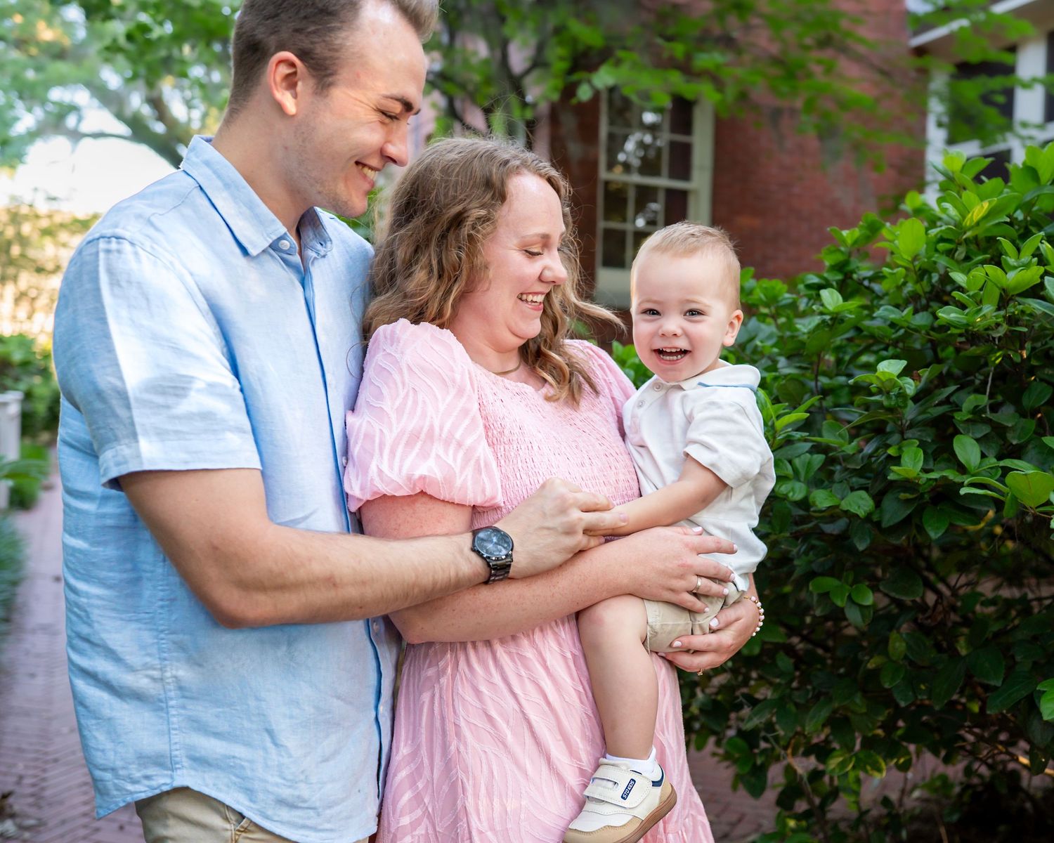 Happy young family with smiling baby boy posing outdoors near brick building and green shrubs.