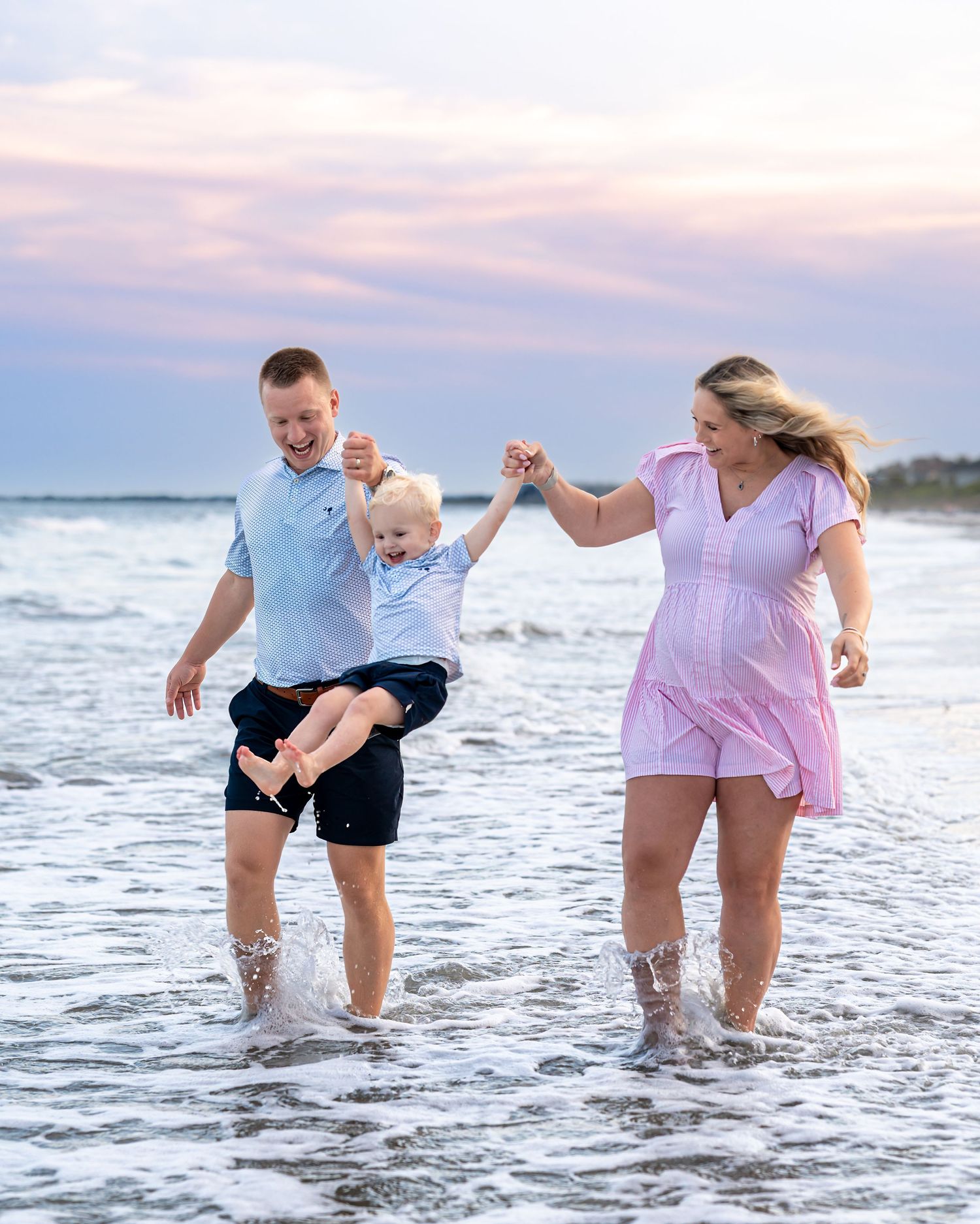 Family of three playing in ocean waves at sunset, parents swinging toddler between them on sandy beach.