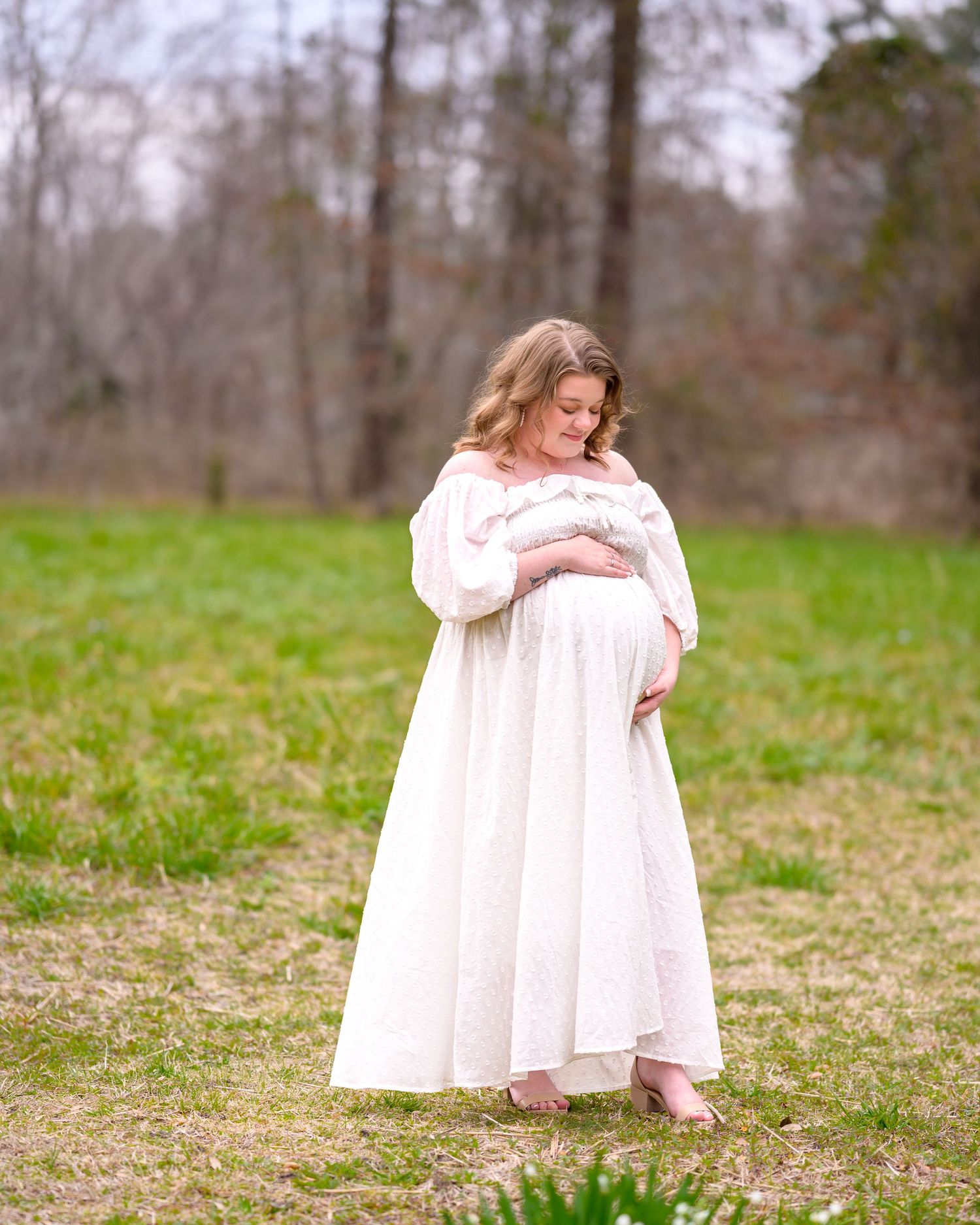 Erin, dressed in a white slowly dress, stands in a large field looking down at and embracing her pregnant belly.