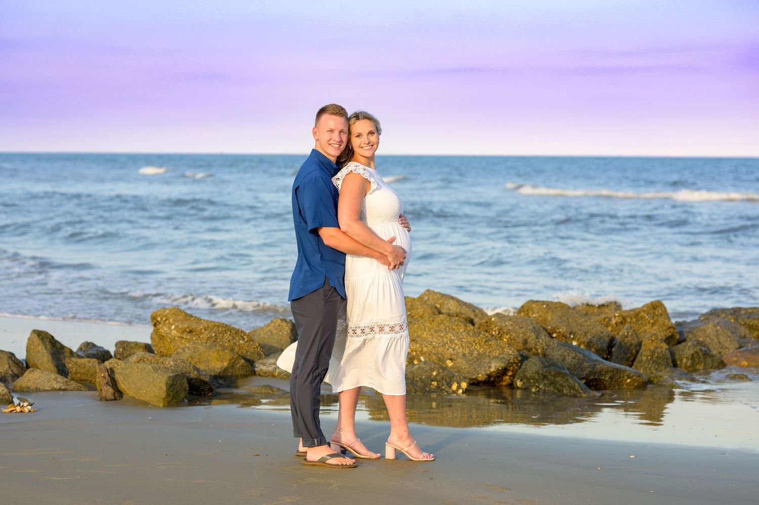 Expecting couple hold each other on the beach, sky with hues of blue and purple behind them