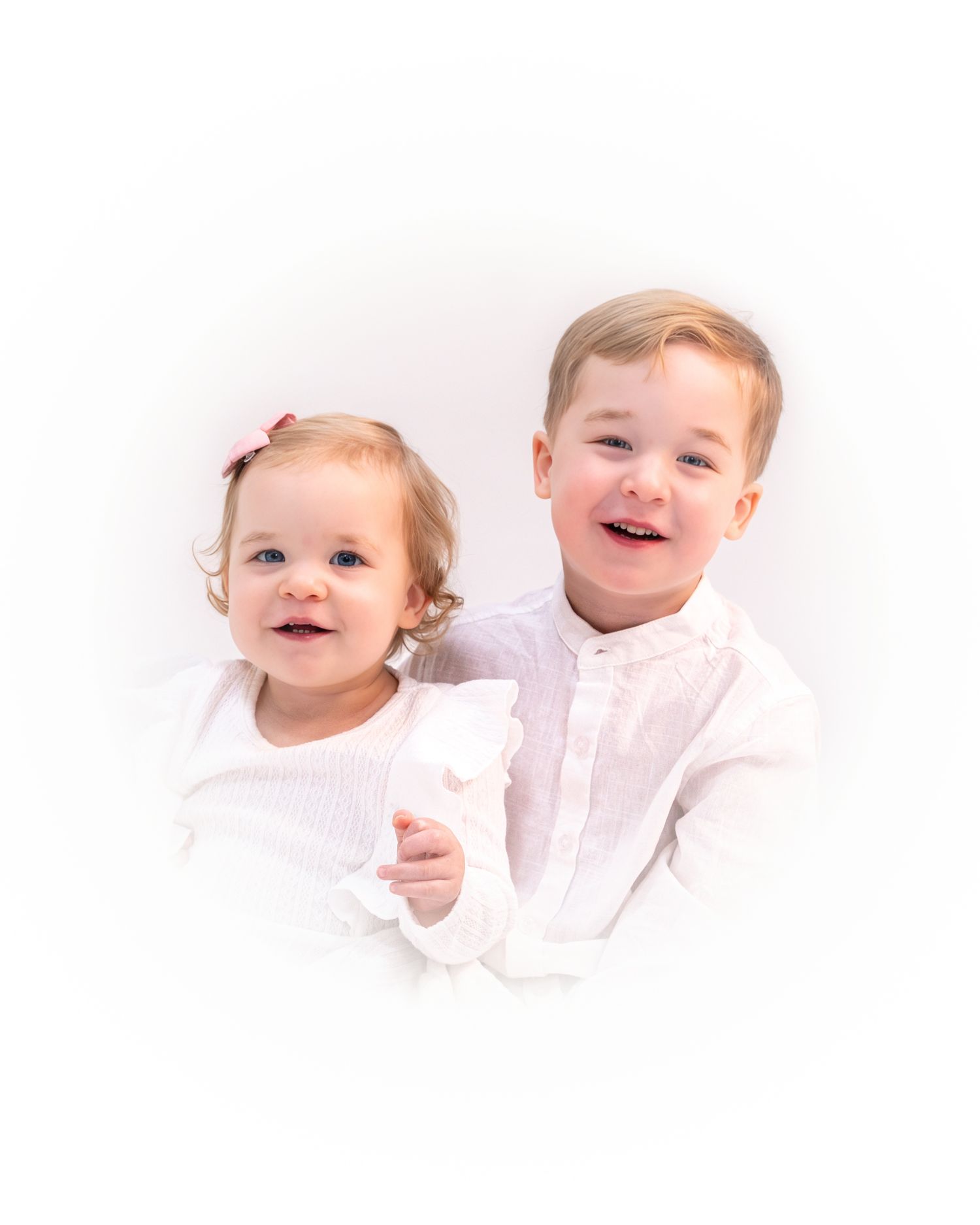 Two young children in white clothing smile joyfully together against a light background.