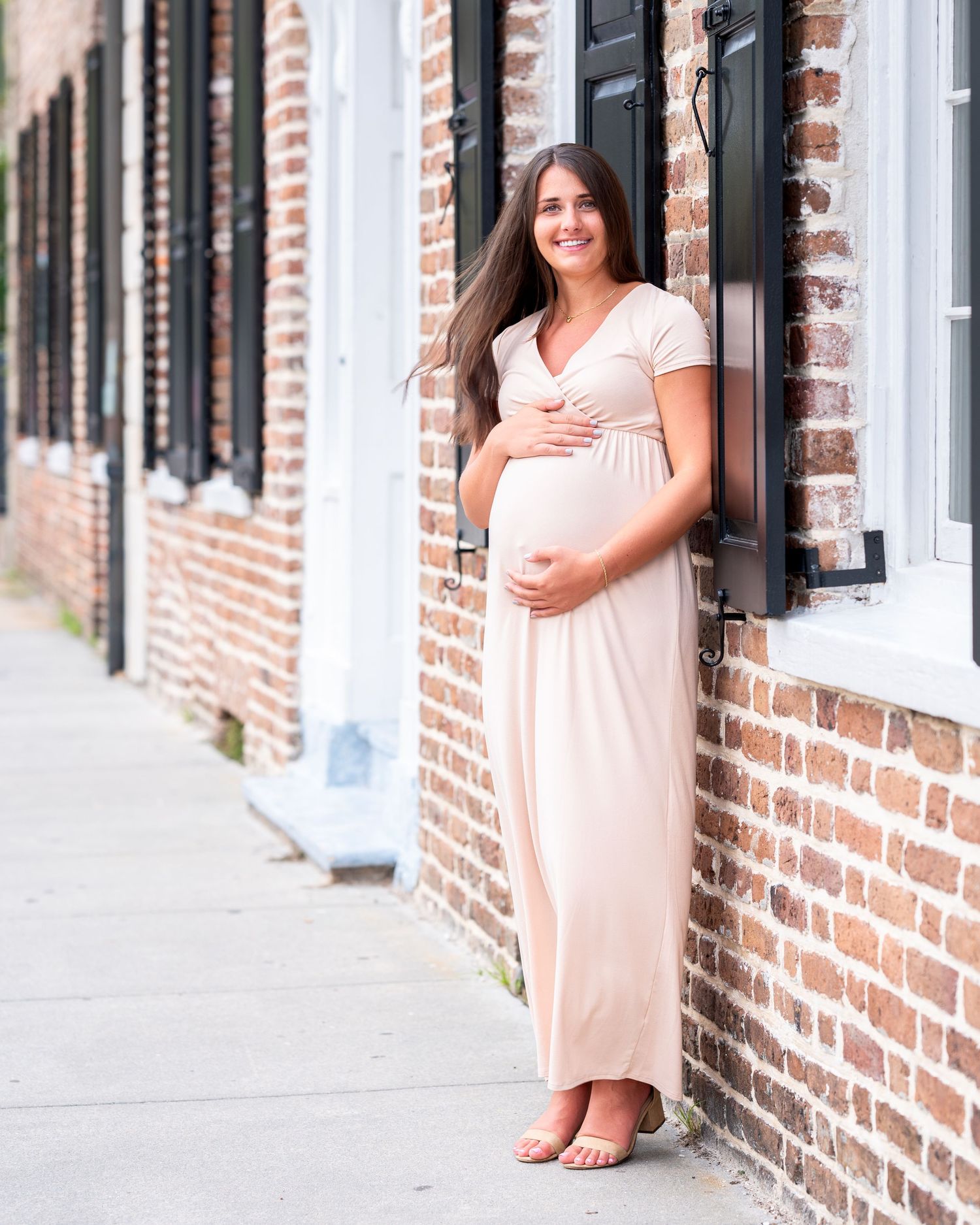 Pregnant woman in white dress stands next to brick wall while holding her bump