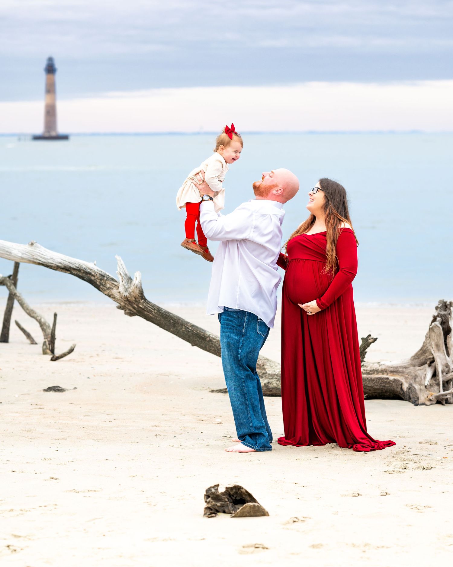 A family shares a tender moment on a beach with a lighthouse visible in the background.