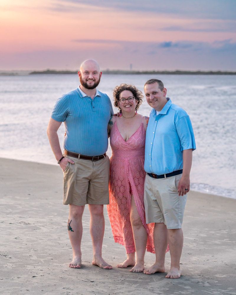 Three people smiling together on a sandy beach at sunset, two men in blue shirts and a woman in a pink dress.