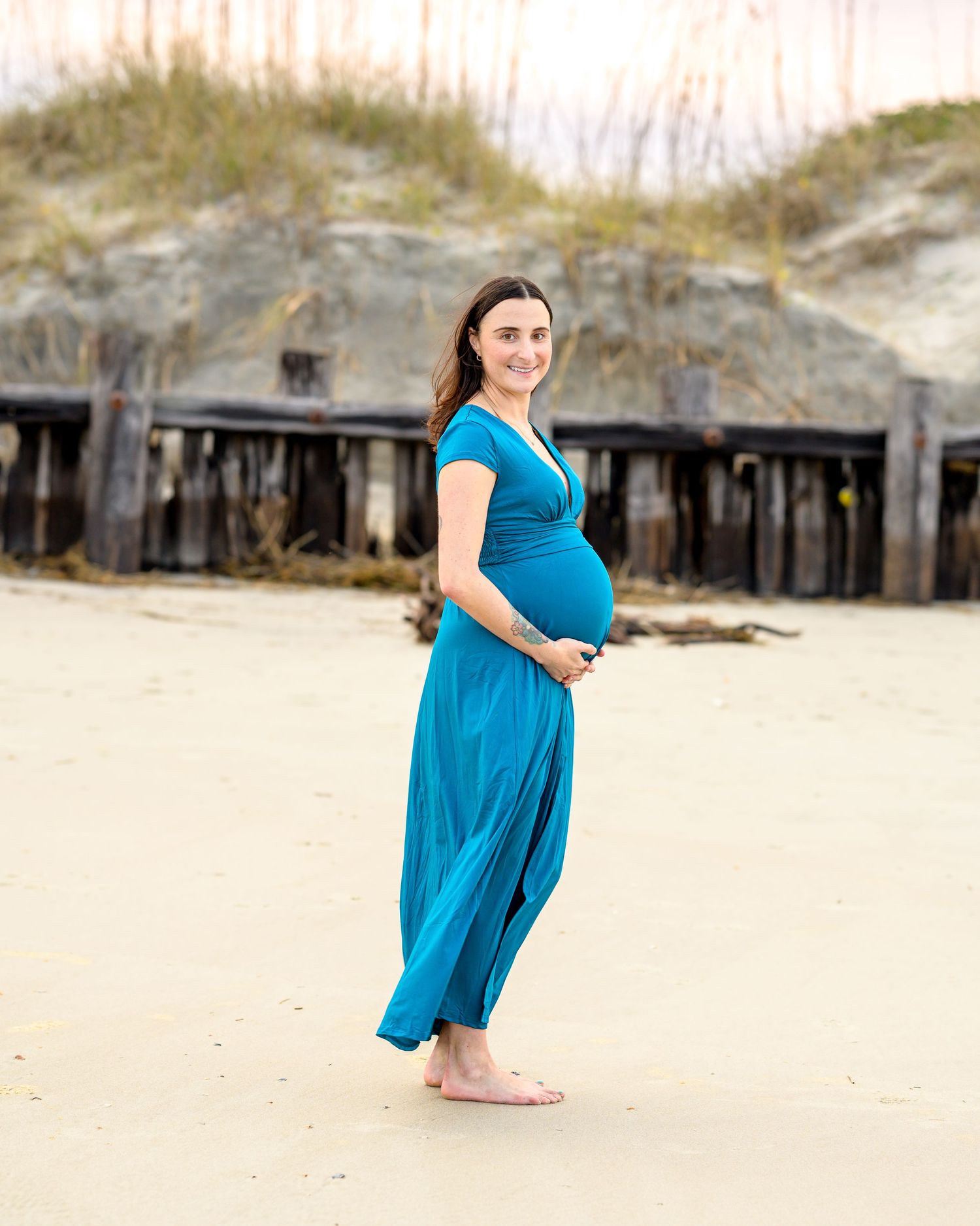 Expecting woman in blue dress holding her baby bump at beach