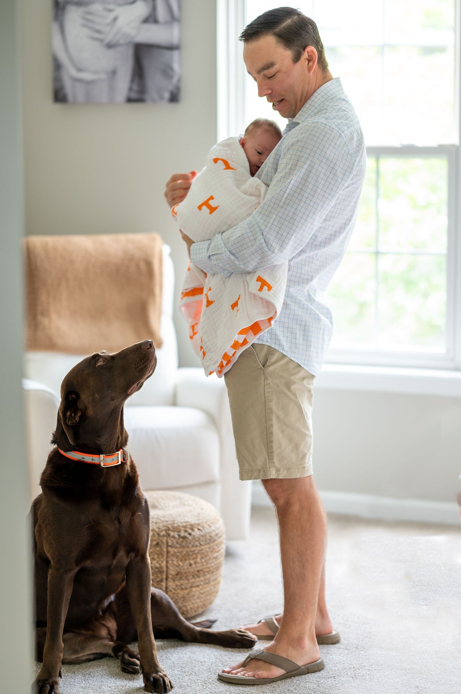 Tom stands in his daughter's nursery holding his newborn while looking down and talking to his chocolate lab.