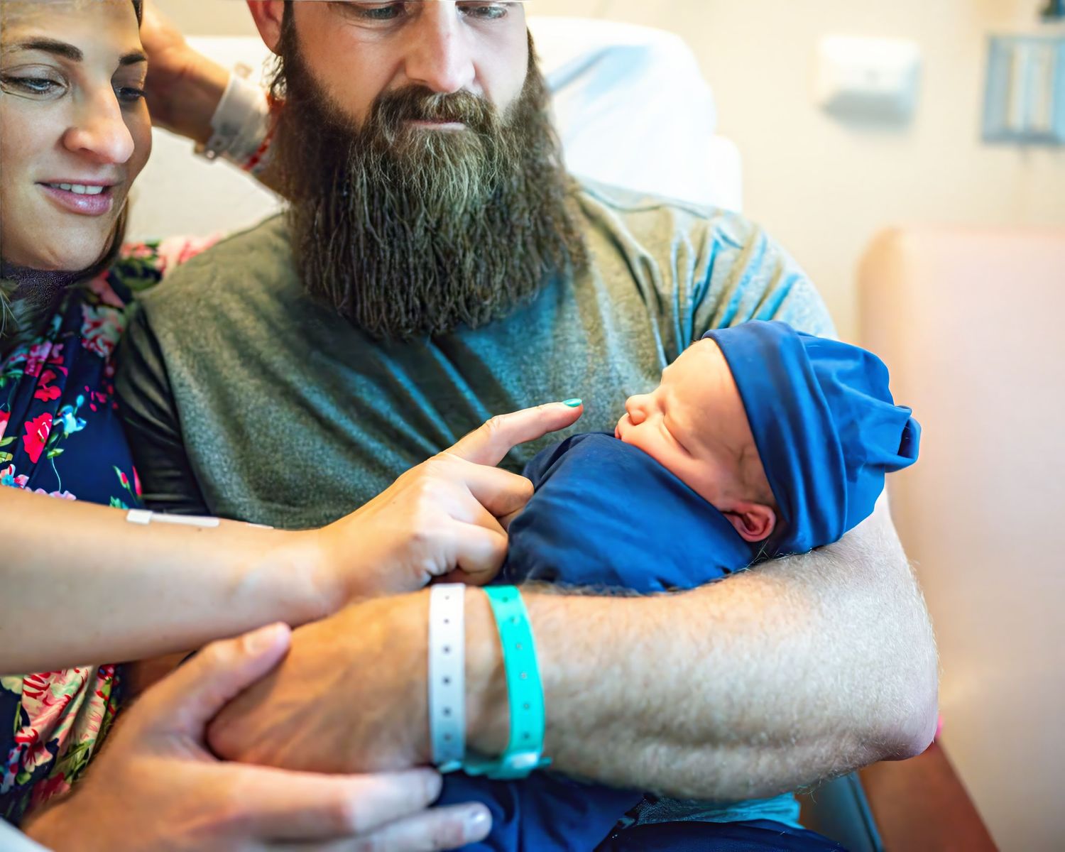 New parents cradle their swaddled newborn baby wearing a blue hat in a hospital room.