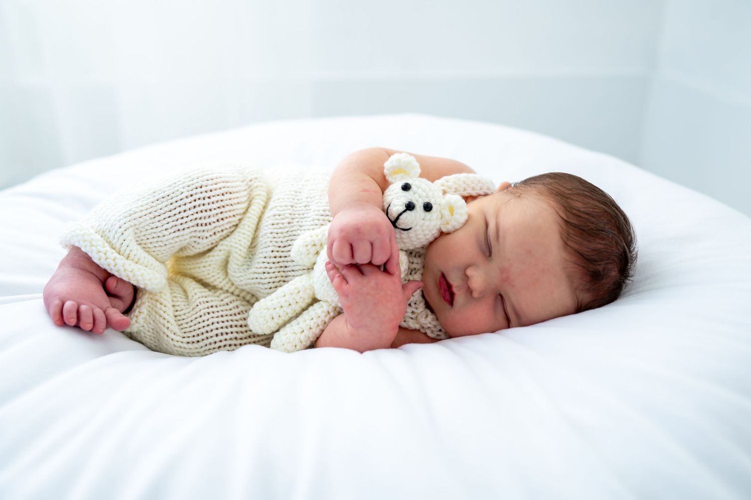 Newborn baby sleeping peacefully in white knit outfit, cuddling a small teddy bear toy on a white bed.