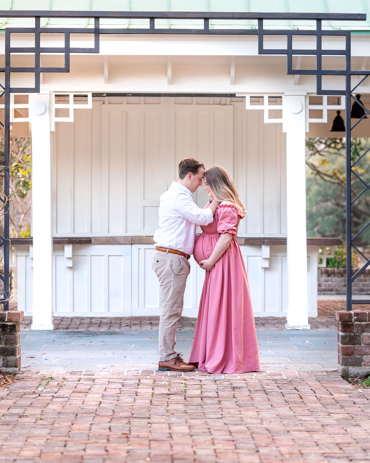 A romantic couple embraces in a white gazebo with red brick flooring, the woman wearing a flowing pink maternity dress.