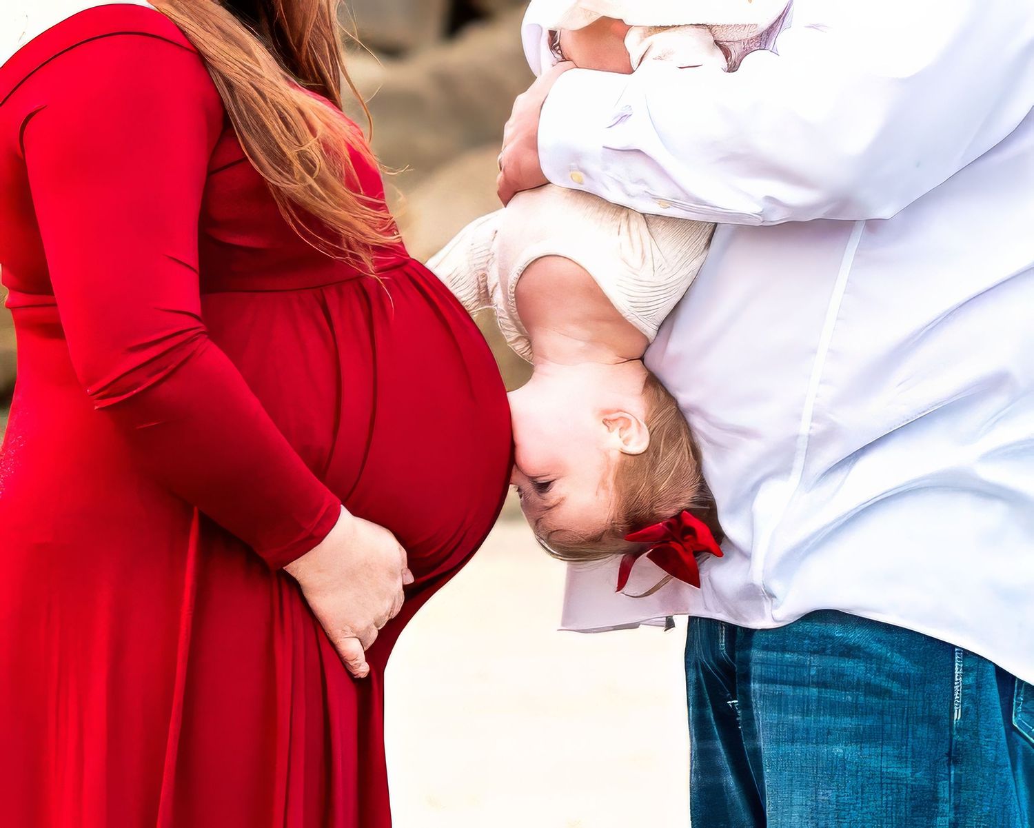 Family portrait of expecting parents in red dress and white shirt playfully posing together outdoors.