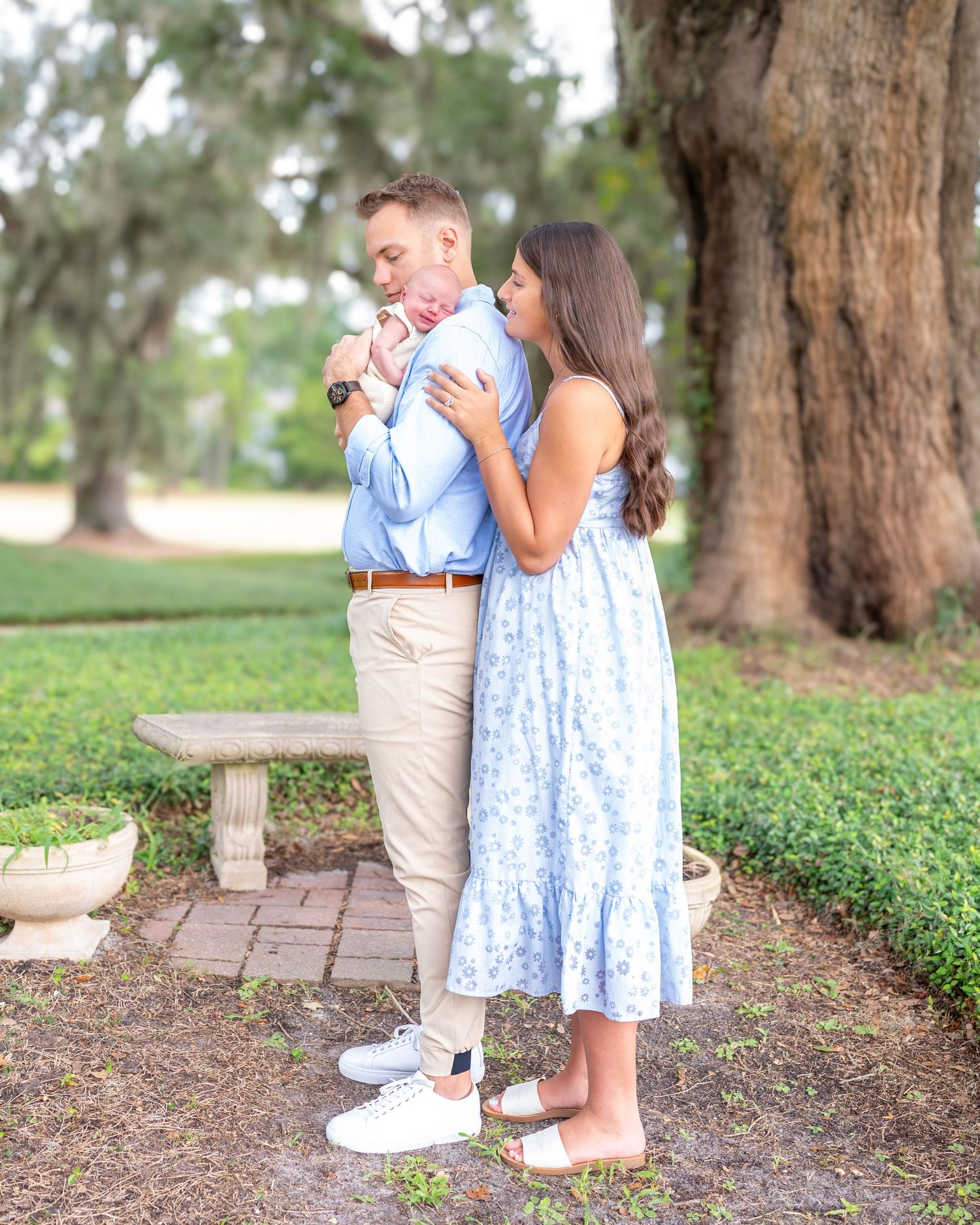 New parents, both dressed in light blue, stand under a tree. Dad holds baby, mom is standing behind dad hugging them