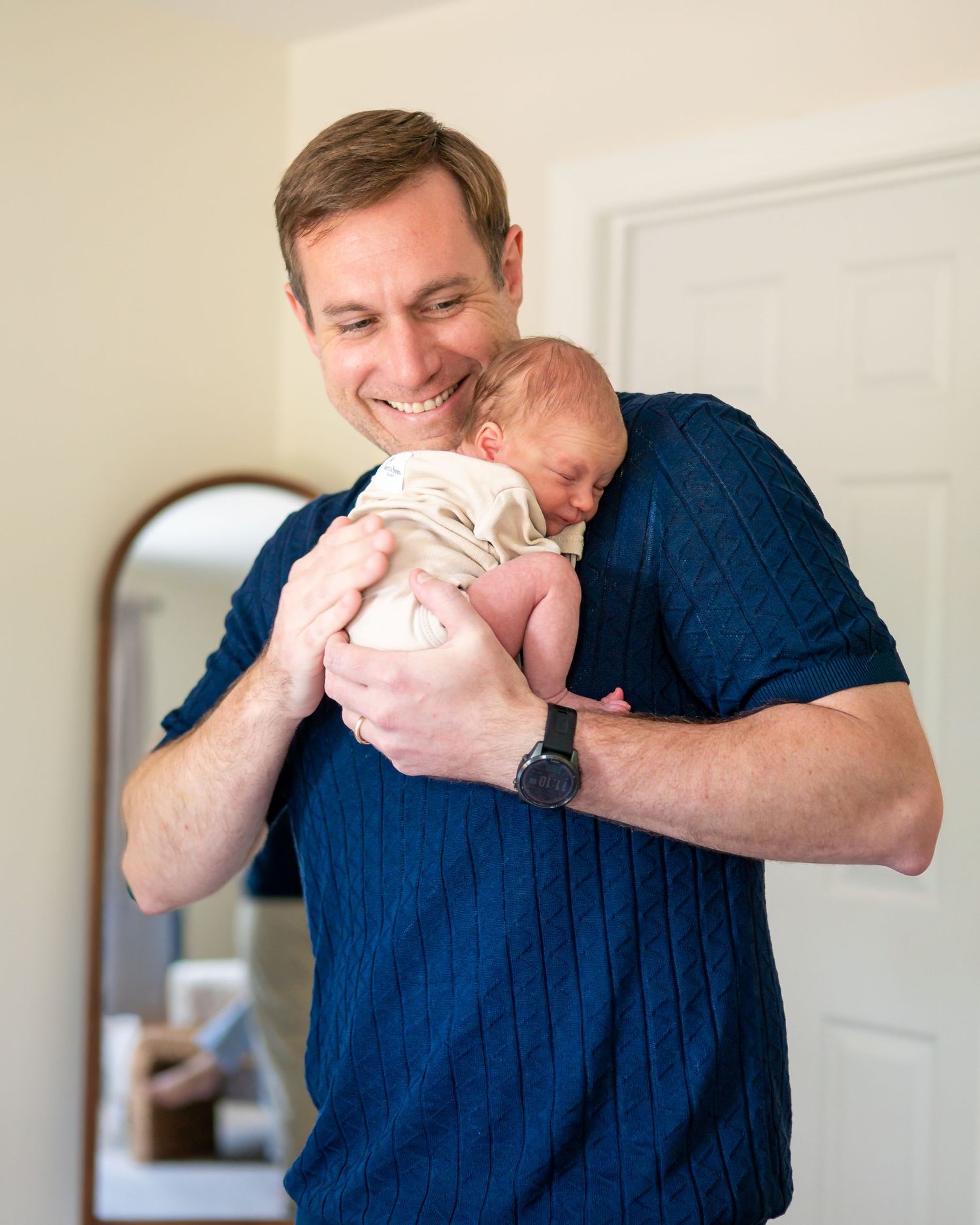 Smiling man in navy sweater tenderly holds sleeping newborn baby close to his chest indoors.