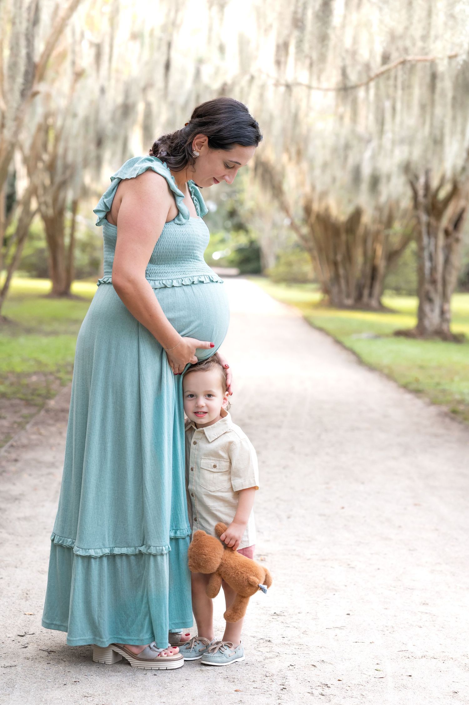 A tender moment captured on a tree-lined path as a pregnant woman in a blue dress stands with a small child.