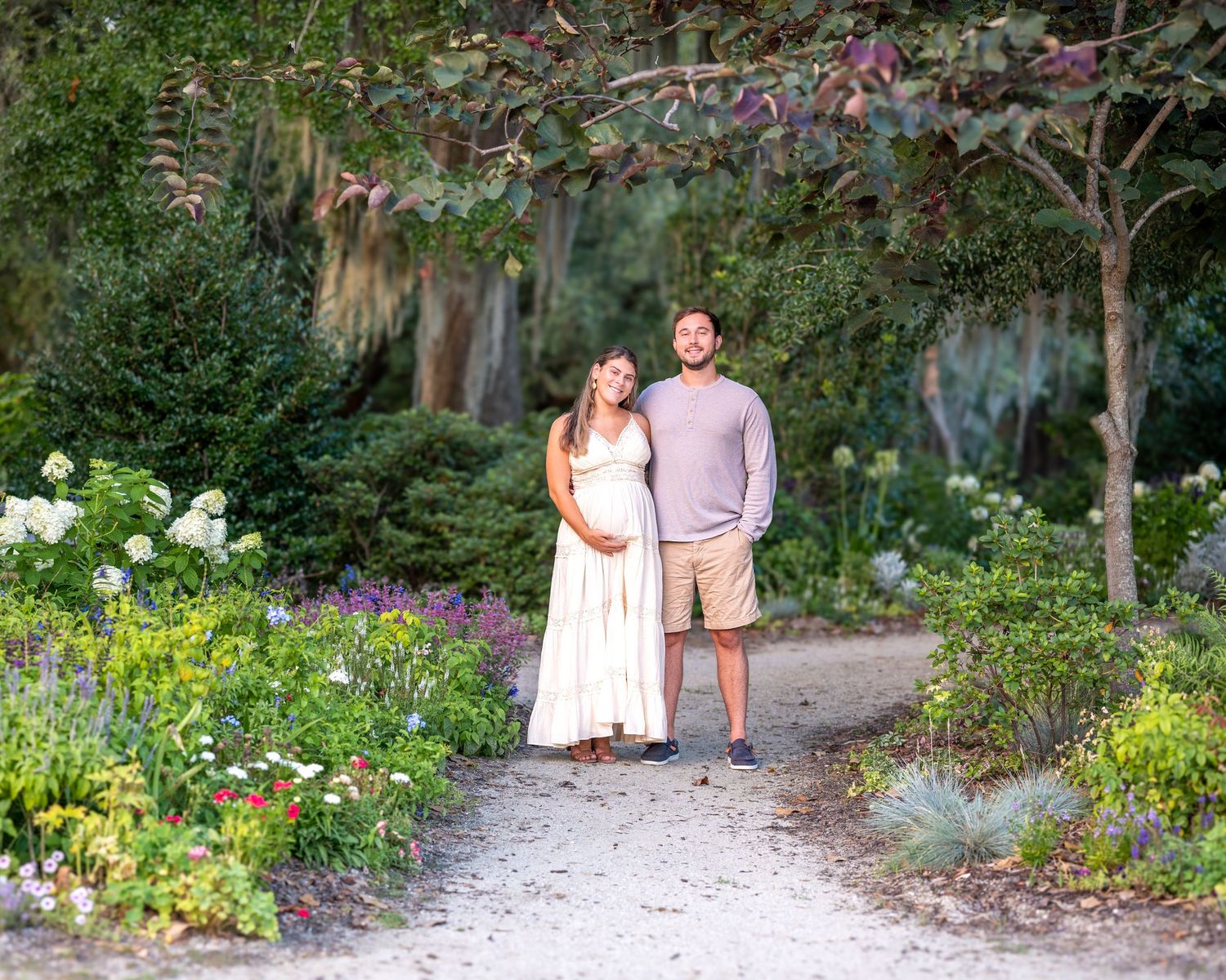 A couple walks along a garden path surrounded by lush flowers and greenery on a sunny day.