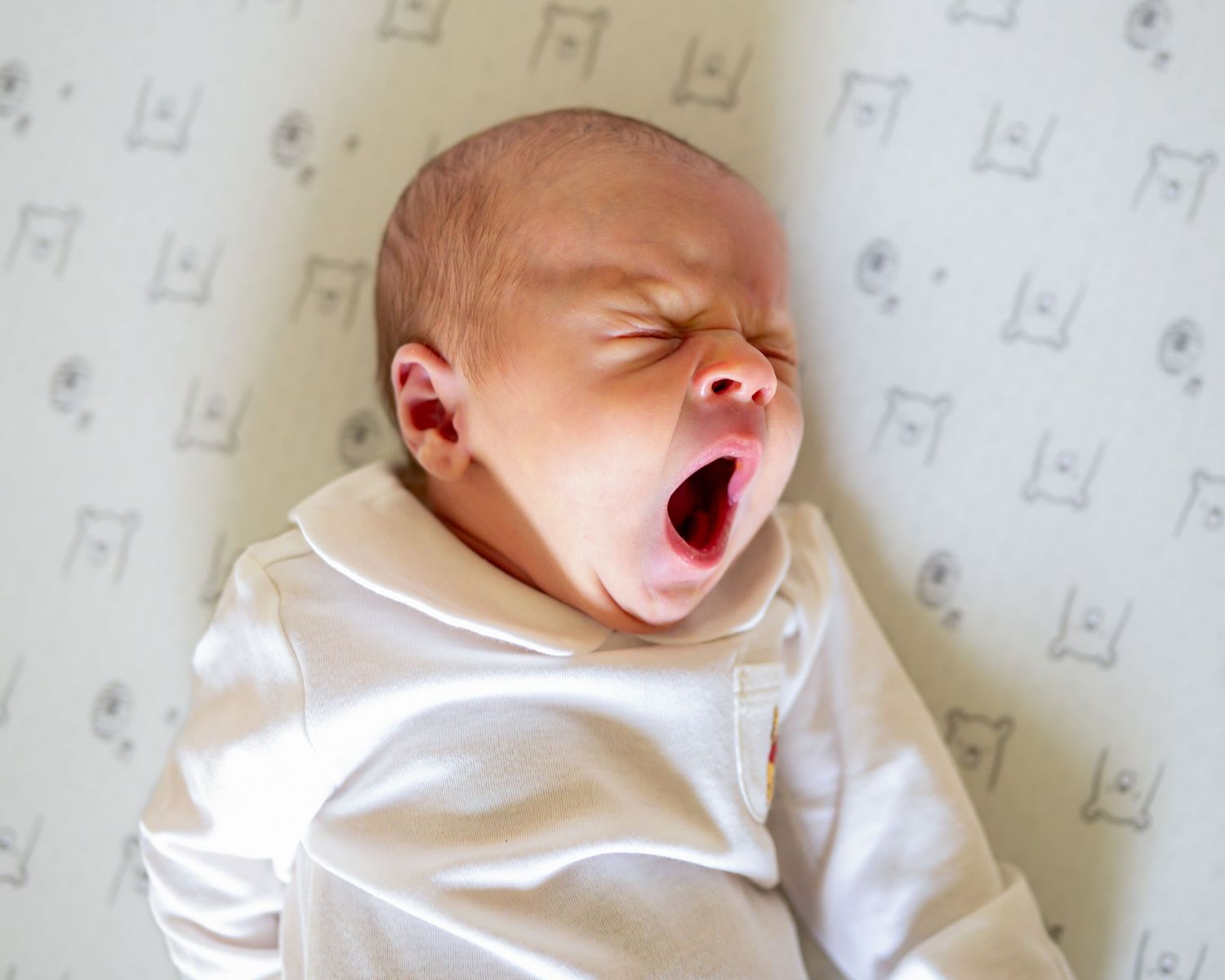 Newborn baby yawning widely while lying on a white patterned blanket, dressed in a white onesie.