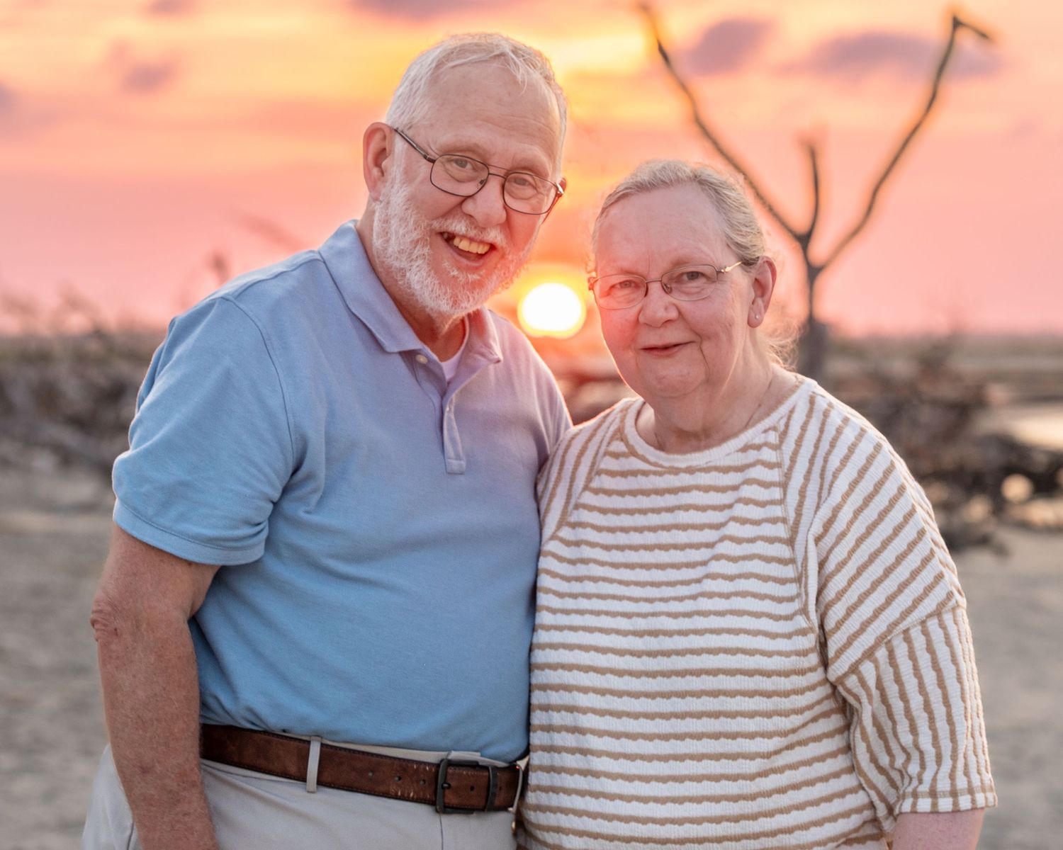 Elderly couple smiling at sunset on a beach, man in blue polo and woman in striped shirt, warm orange sky.