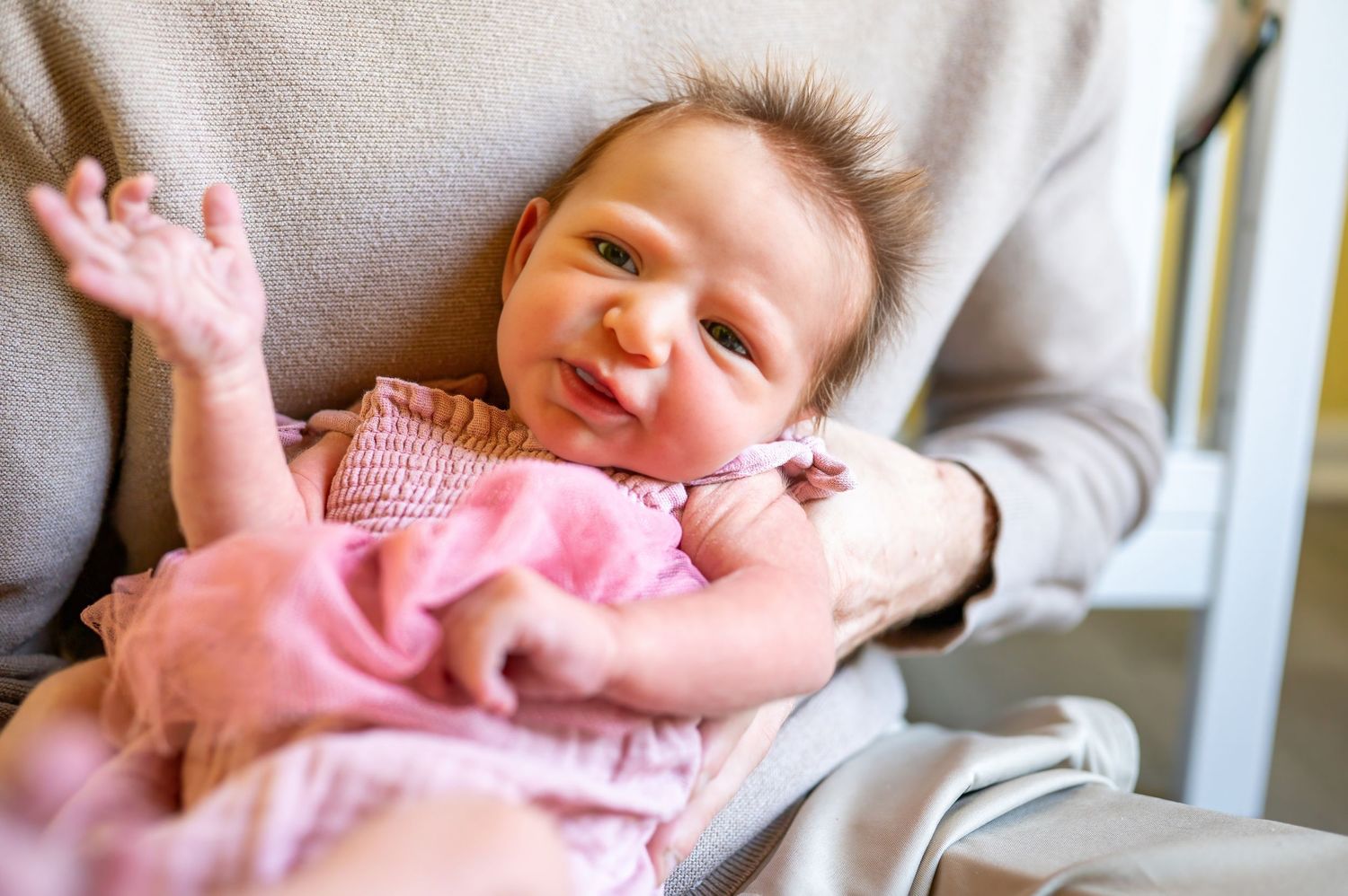 Newborn girl is wearing pink dress lies on her father's chest with arm raised while smiling joyfully.