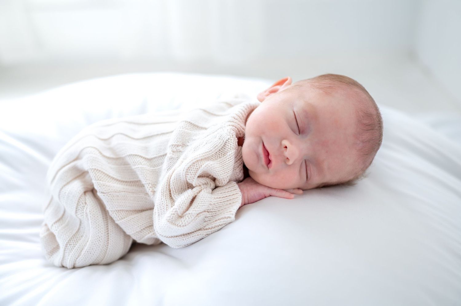 Peaceful newborn baby sleeping swaddled in a cream knit blanket on white bedding in soft natural light.