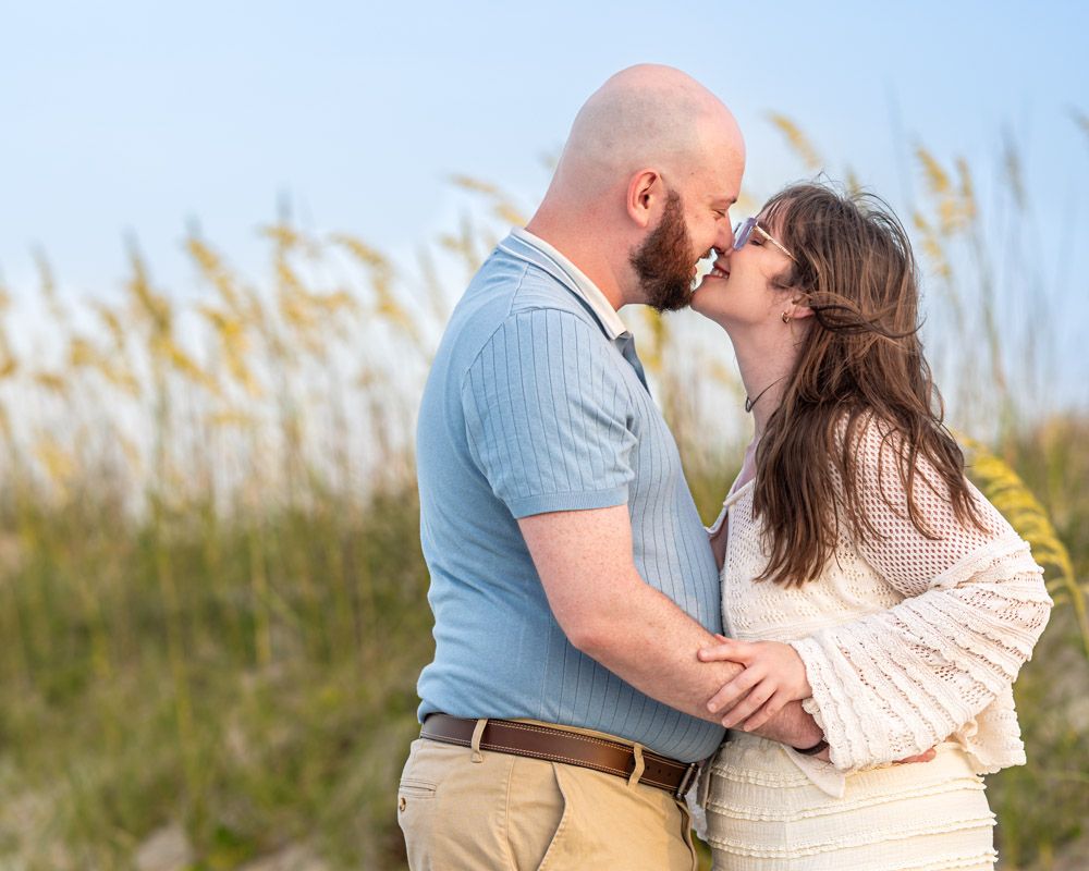 Couple sharing a romantic kiss on a beach with sea oats in the background on a sunny day.