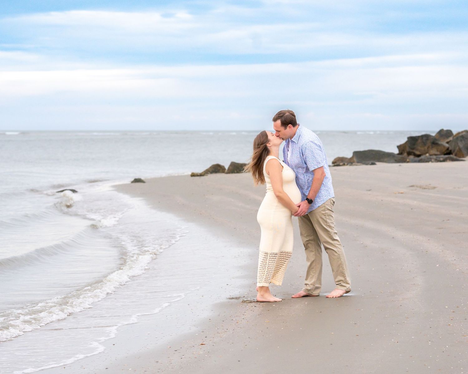 Couple sharing a romantic kiss on a sandy beach with gentle ocean waves and rocky shoreline in the background during their maternity session