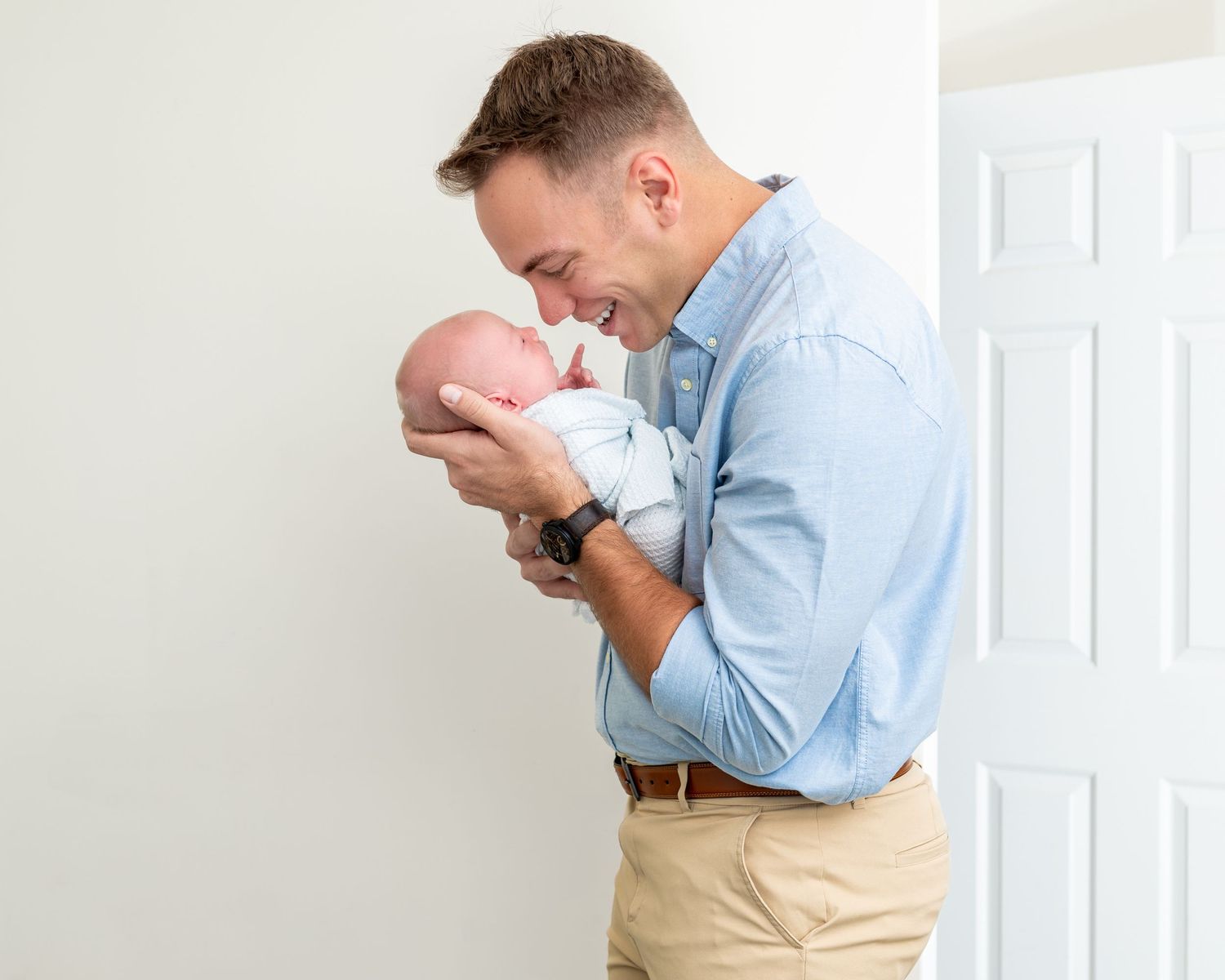 Dad wearing blue shirt with rolled sleeves holds his newborn son so that they are nose to nose
