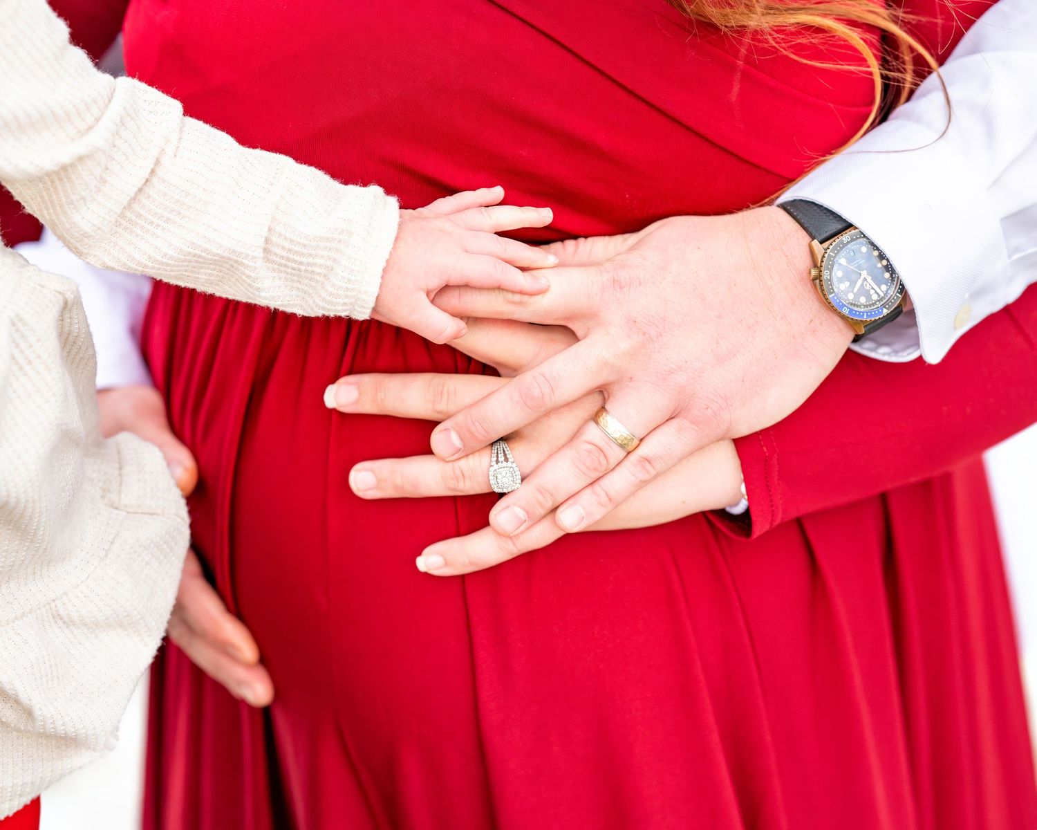 Two hands rest gently on a red maternity dress, one wearing a watch and one in a cream colored sweater.