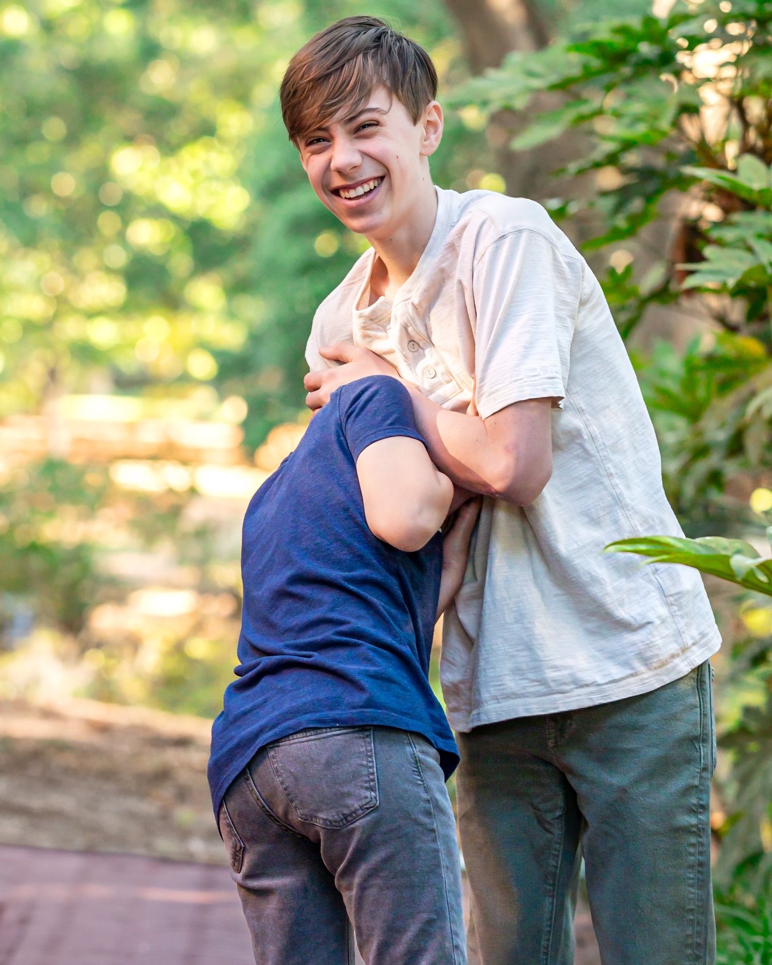 Two young men laughing and playfully wrestling outdoors on a sunny day surrounded by green trees.