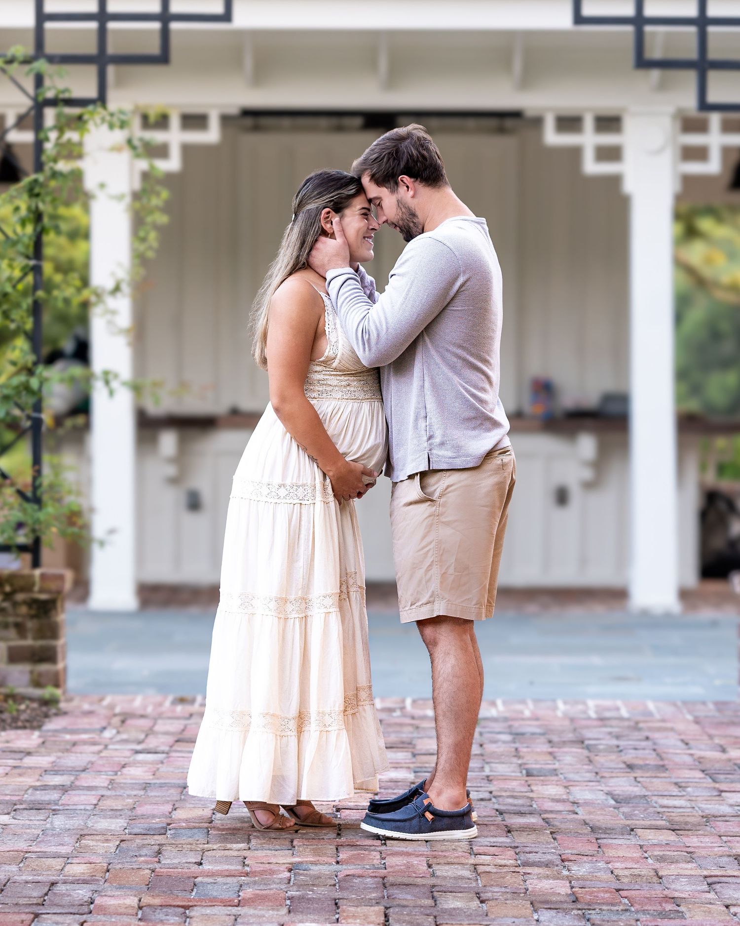 Sweet romantic couple in light summer outfits sharing an intimate moment under a white gazebo on brick patio during their maternity session