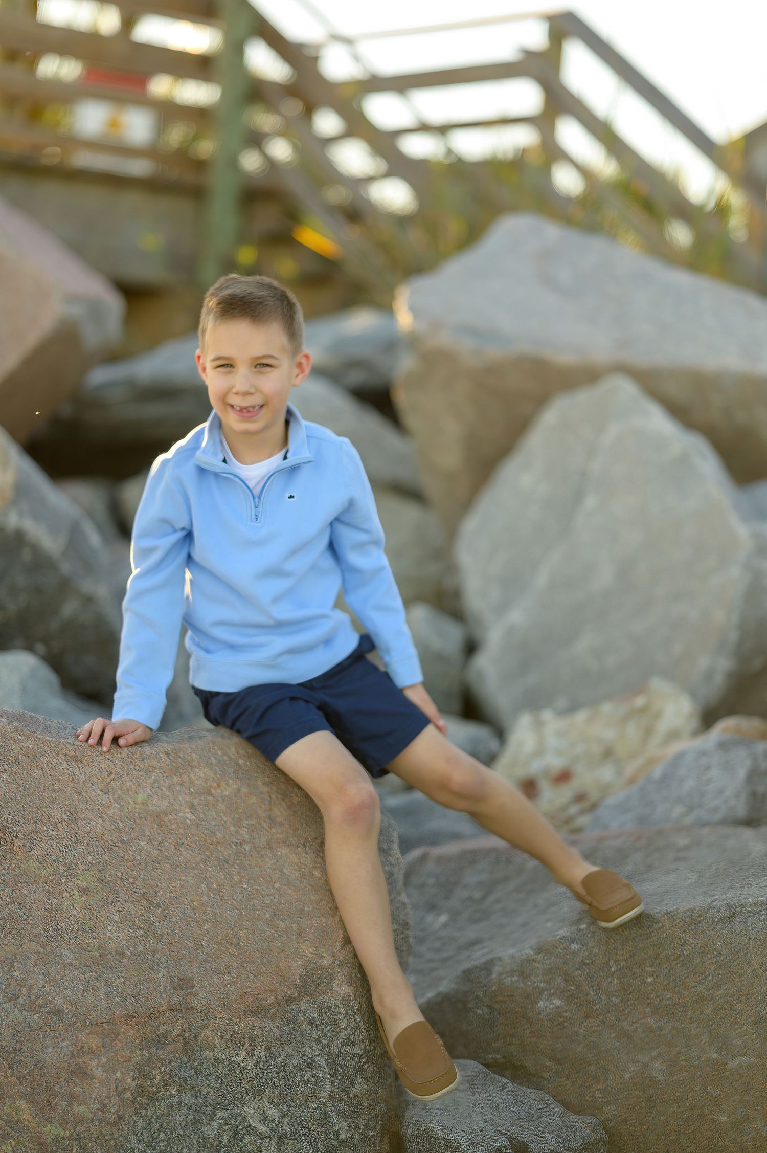 Young boy with blue long sleeve shirt sitting on rocks at the beach
