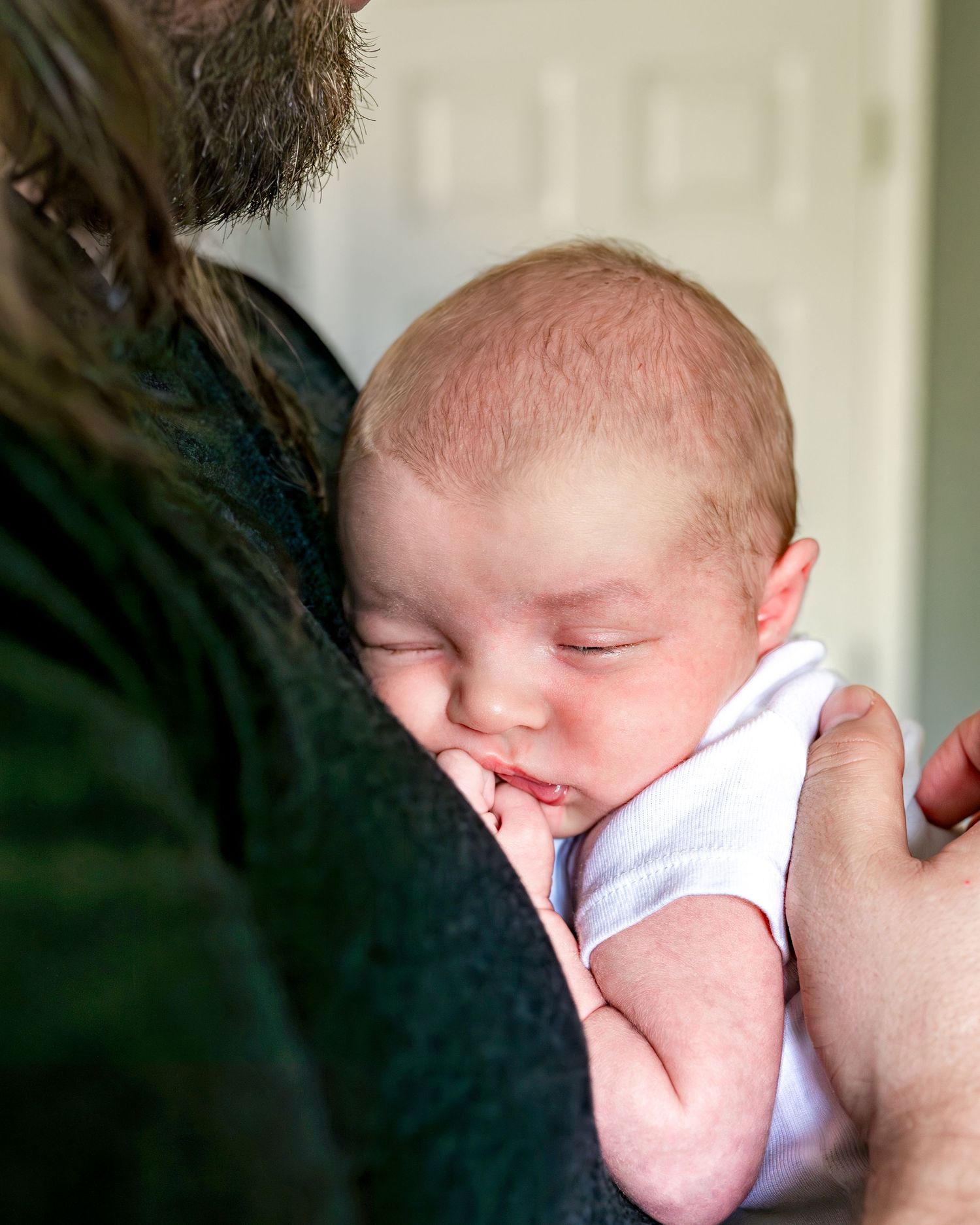 A sleeping newborn baby dressed in white rests peacefully against someone in dark clothing.