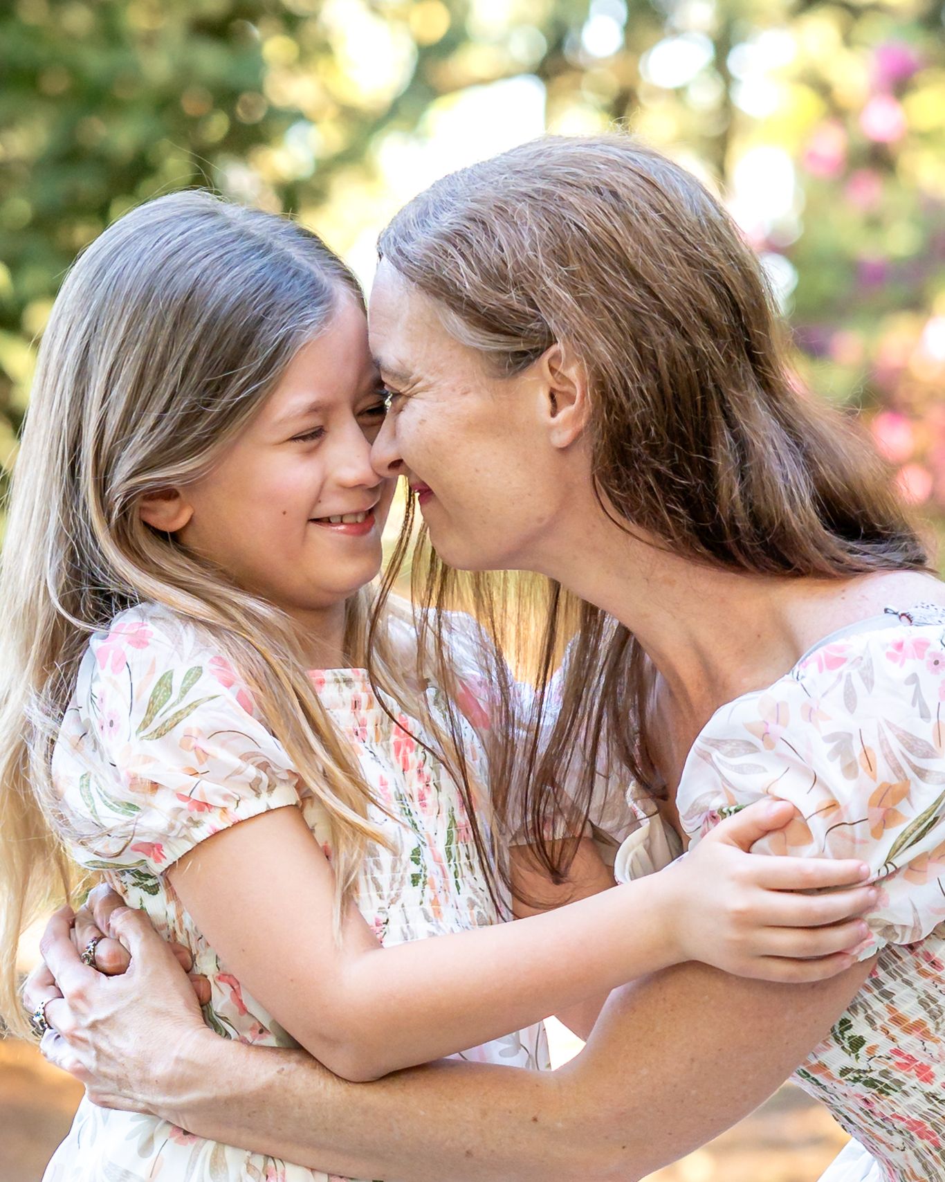 A mother and young daughter in matching floral dresses share a tender nose-to-nose moment outdoors.