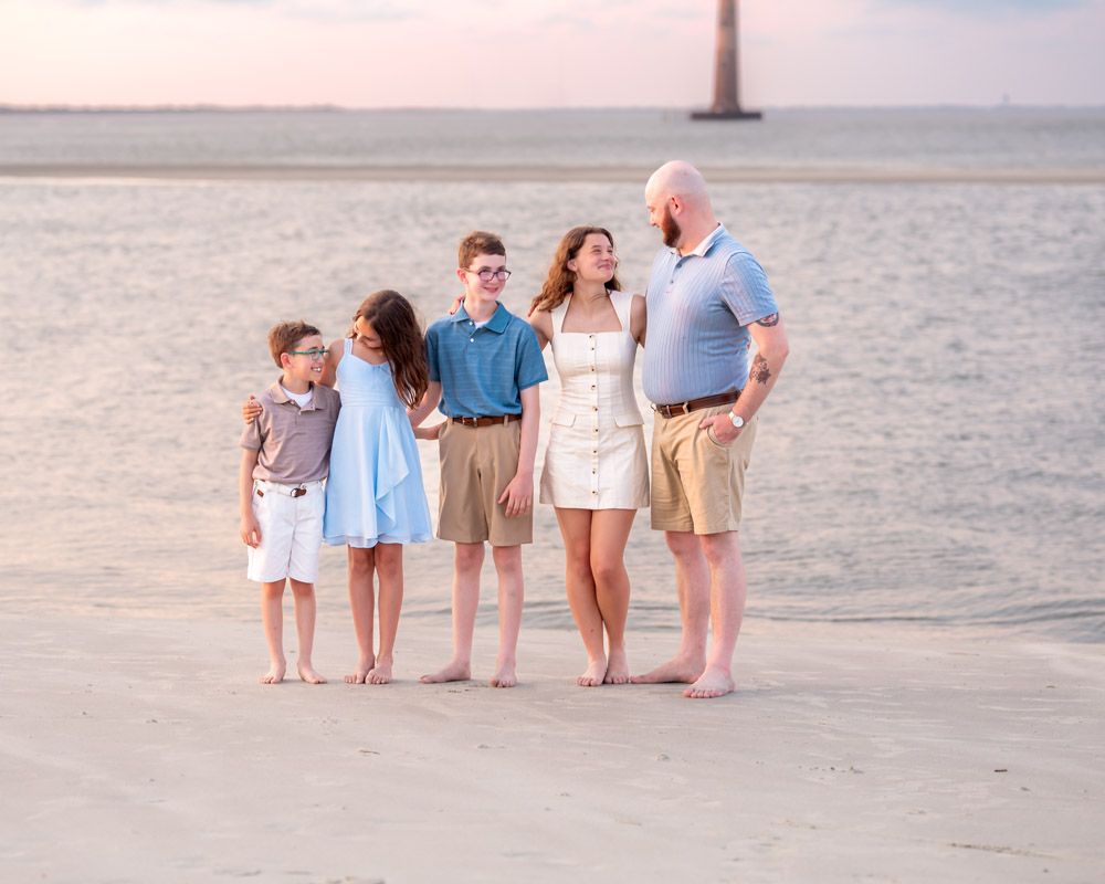 Family of five posing barefoot on sandy beach at sunset with lighthouse in background.