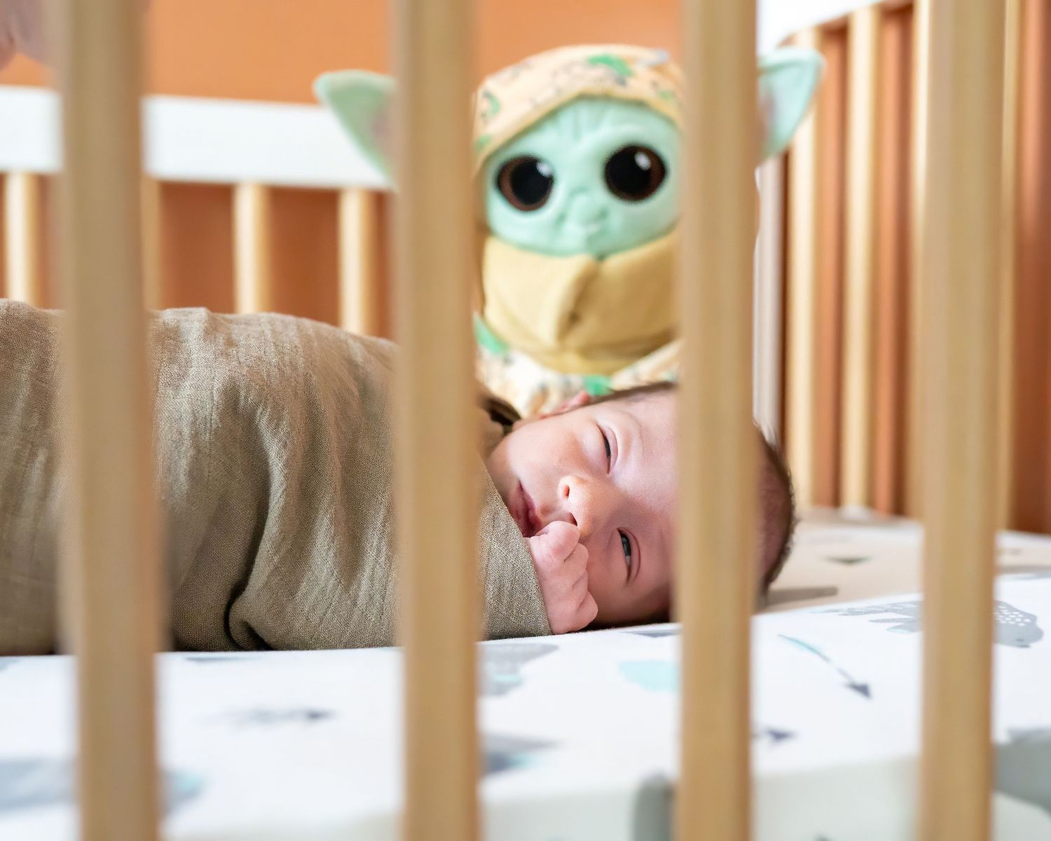 Baby lying in crib with stuffed baby Yoda toy looking up through wooden slats.